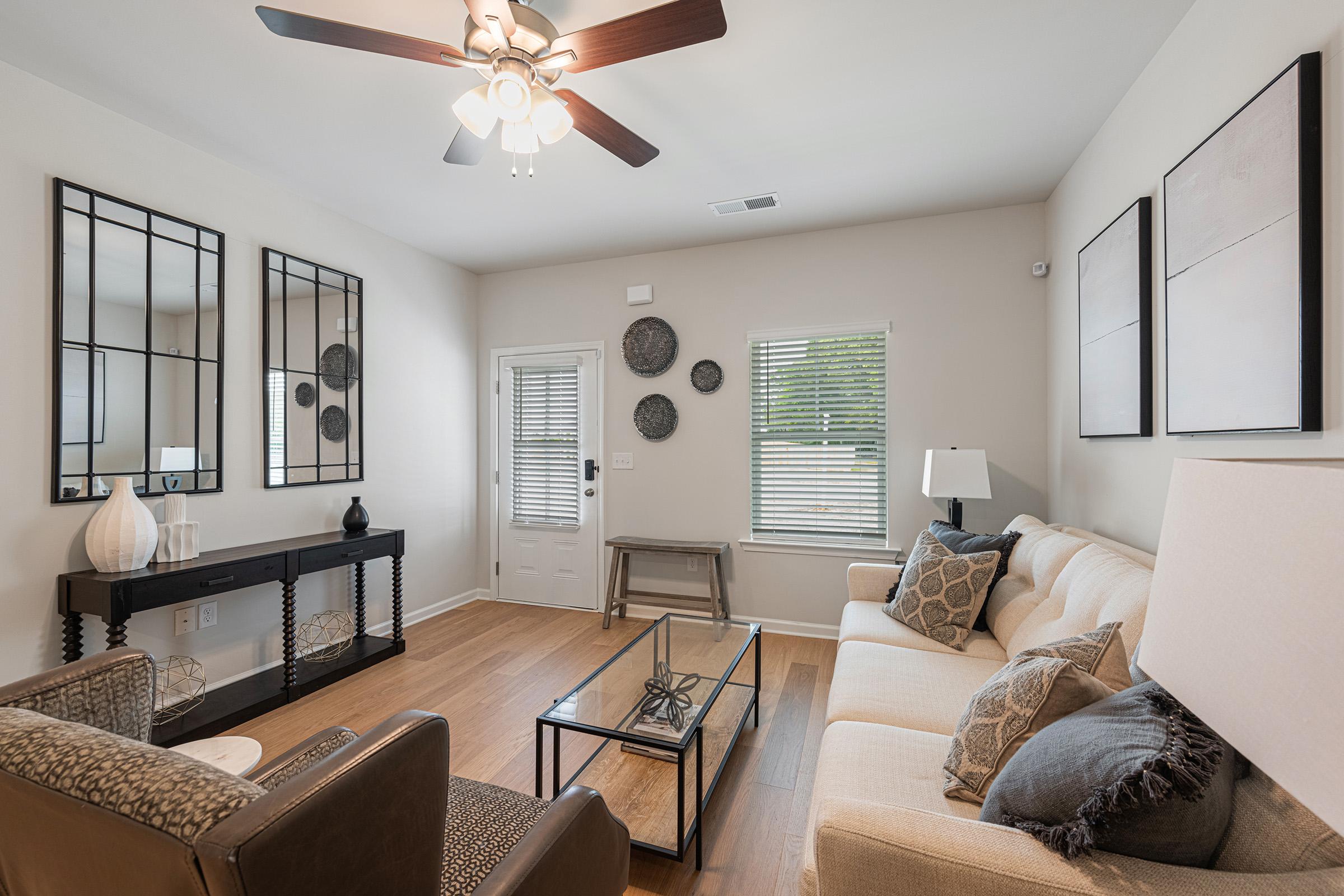 A modern living room featuring a beige sofa with decorative cushions, a glass coffee table, and a stylish console table. The room has decorative mirrors on the wall, round wall art pieces, and a ceiling fan. Natural light streams in through a window, creating a warm and inviting atmosphere.