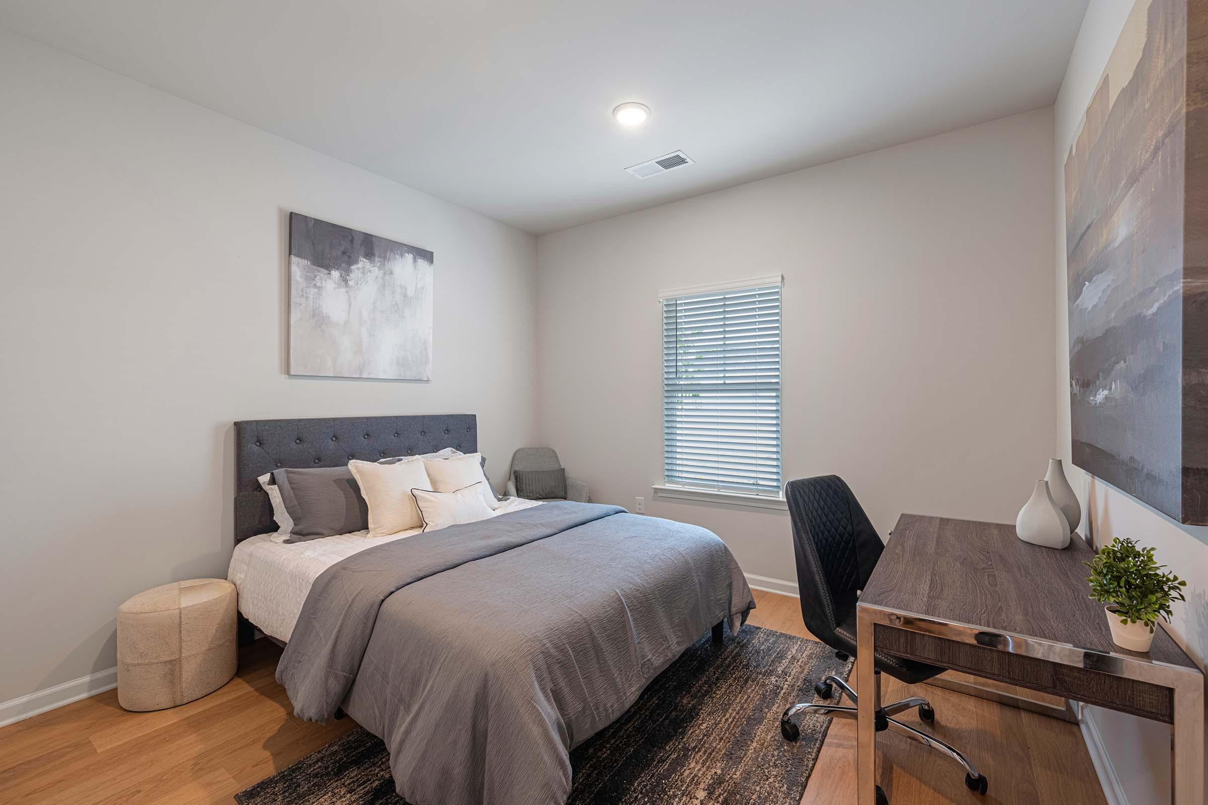 A modern bedroom featuring a gray upholstered bed with a neatly arranged duvet and pillows. There's a small desk with a chair against the wall, a potted plant, and artwork above the bed. Natural light streams through a window with blinds, and the floor is wooden. The overall decor is minimalist and soothing.