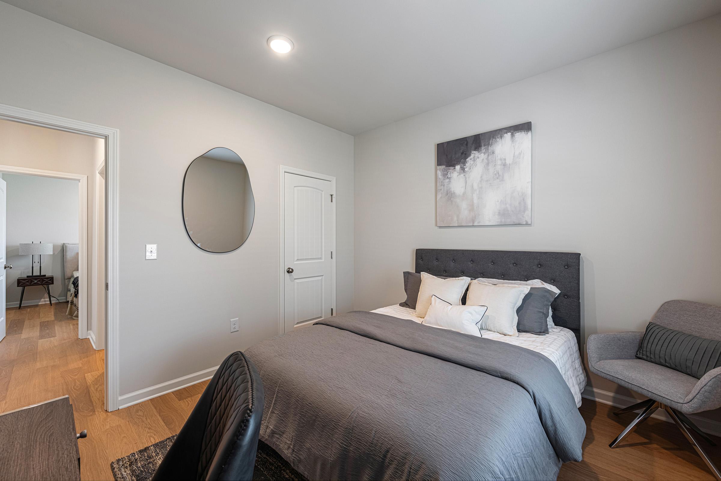 A modern, minimalist bedroom featuring a gray bed with decorative pillows, a round mirror on the wall, and a small armchair. The walls are painted in a light color, and there is a door leading to another room visible in the background. The flooring is hardwood, adding warmth to the space.