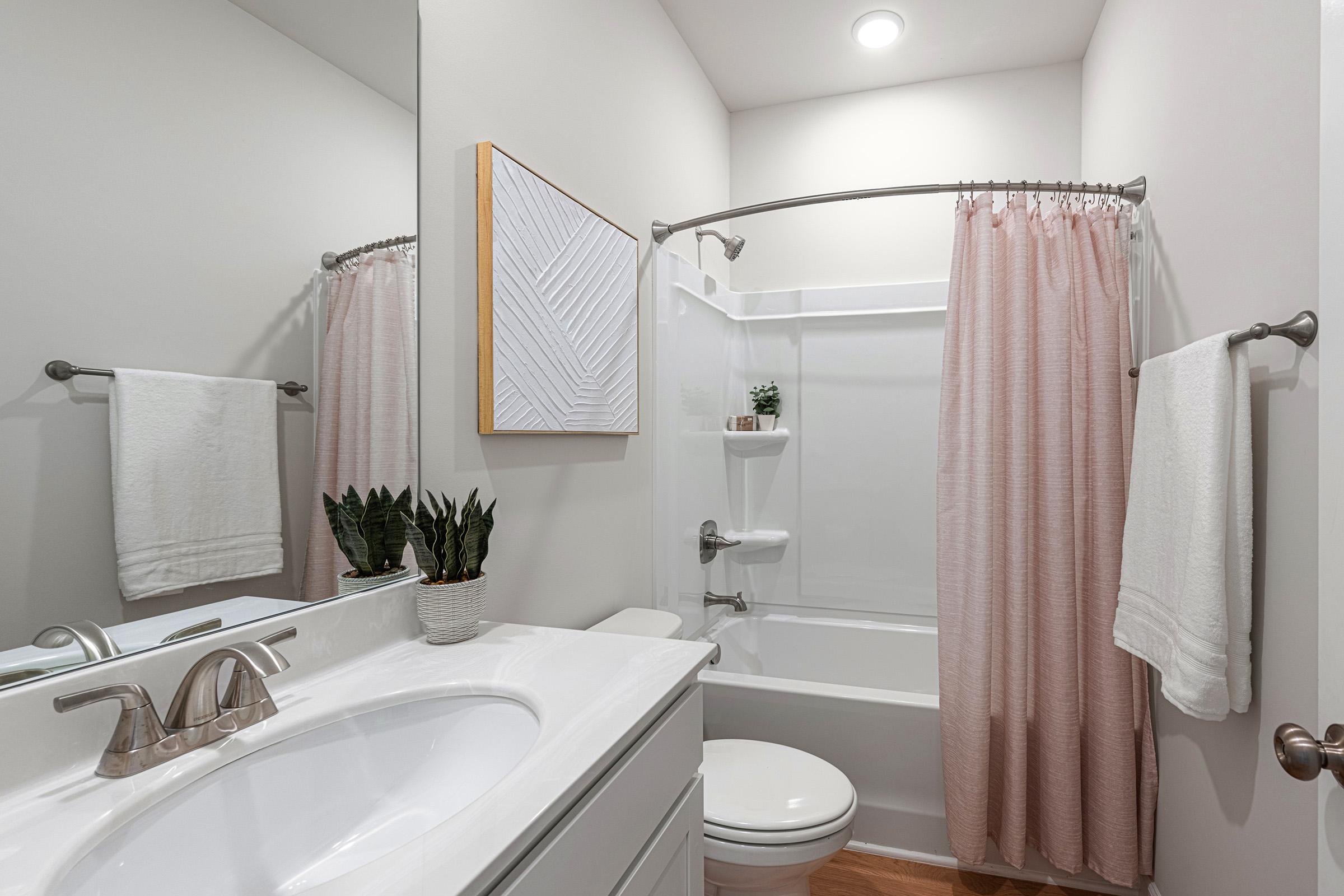 A modern bathroom featuring a white sink, large mirror, and a bathtub with a pink shower curtain. Two white towels hang on towel bars, and a small potted plant decorates the counter. The walls are painted in neutral tones, creating a bright and clean atmosphere.
