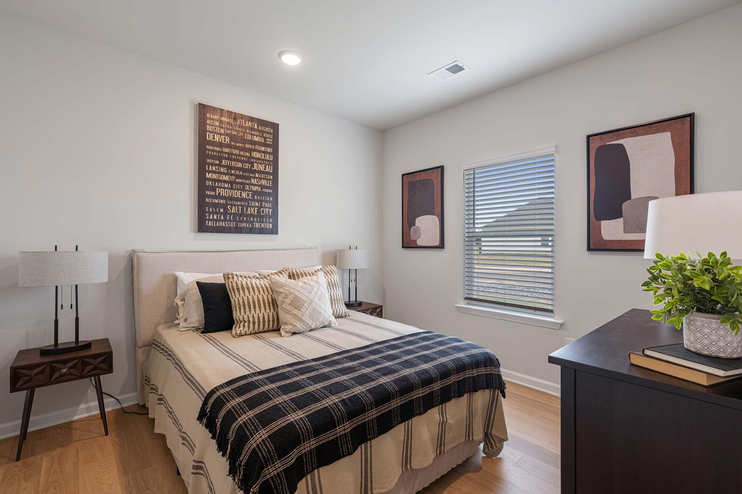 A cozy bedroom featuring a bed with a plaid blanket, two bedside tables with lamps, a modern dresser, and wall art. The walls are painted white, and there is a window allowing natural light to enter. The room has a minimalist and inviting decor.