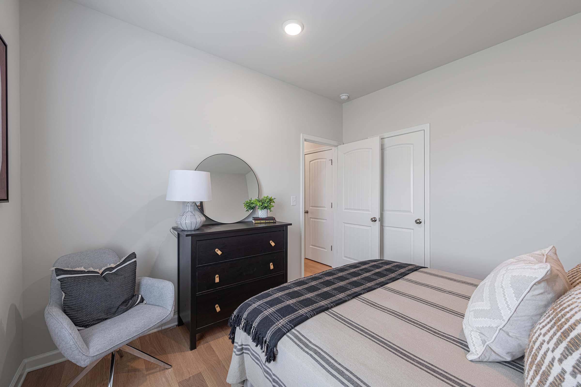A cozy bedroom featuring a bed with a patterned throw, a black dresser with gold handles, a round mirror on the dresser, a grey accent chair in the corner, and light-colored walls. The room has a warm and inviting atmosphere, with natural light coming from the ceiling fixture.