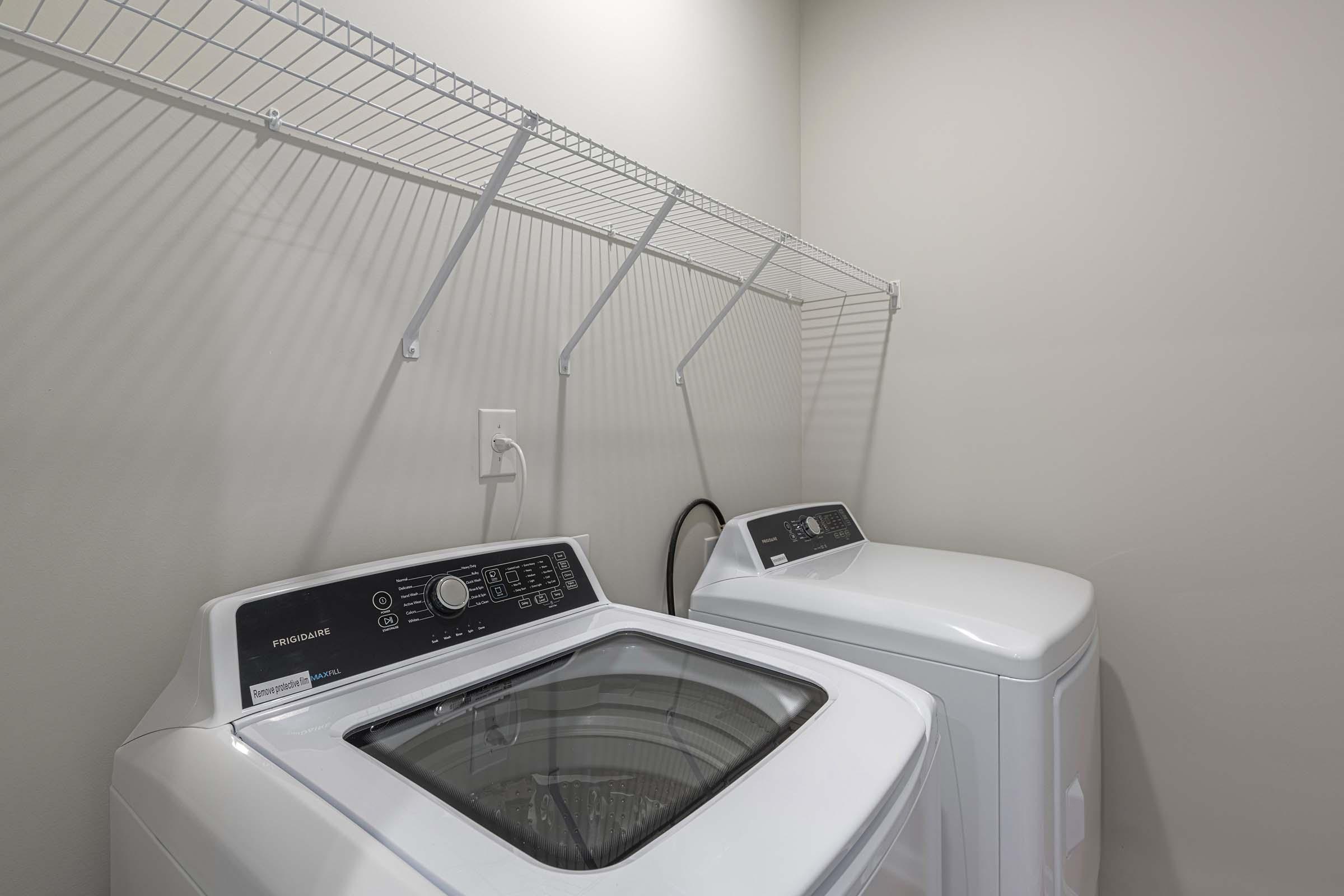 A bright laundry room featuring a modern washing machine and dryer side by side. Above, there is a metal wire shelf mounted on the wall. The walls are painted a light color, creating a clean and organized space ideal for laundry chores.