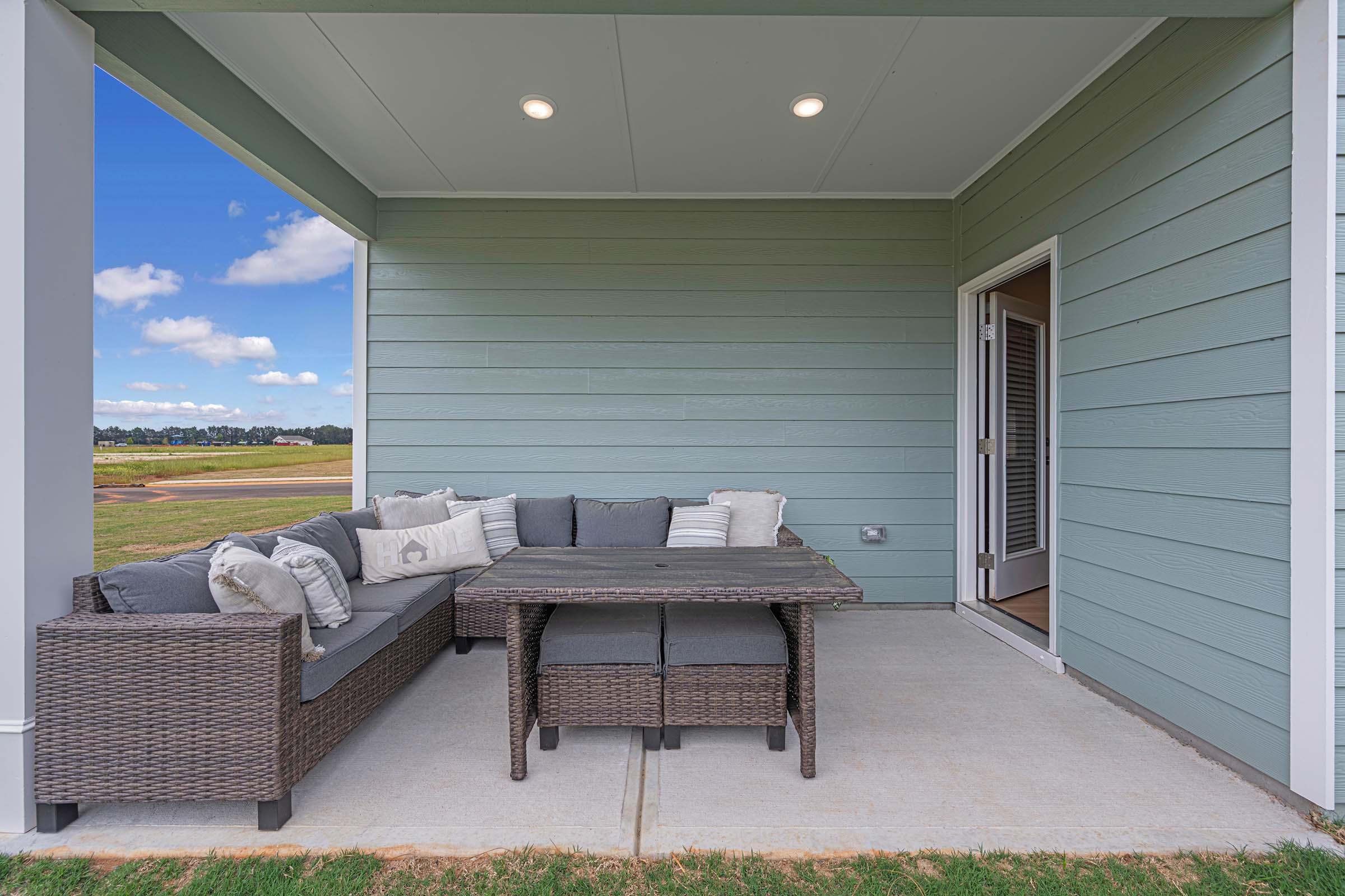 A cozy outdoor seating area with a large gray sectional sofa adorned with decorative pillows. In front of the sofa is a dark wicker coffee table. The space is shaded by a roof and has light green walls. In the background, there is a view of a grassy field under a clear blue sky with a few clouds.