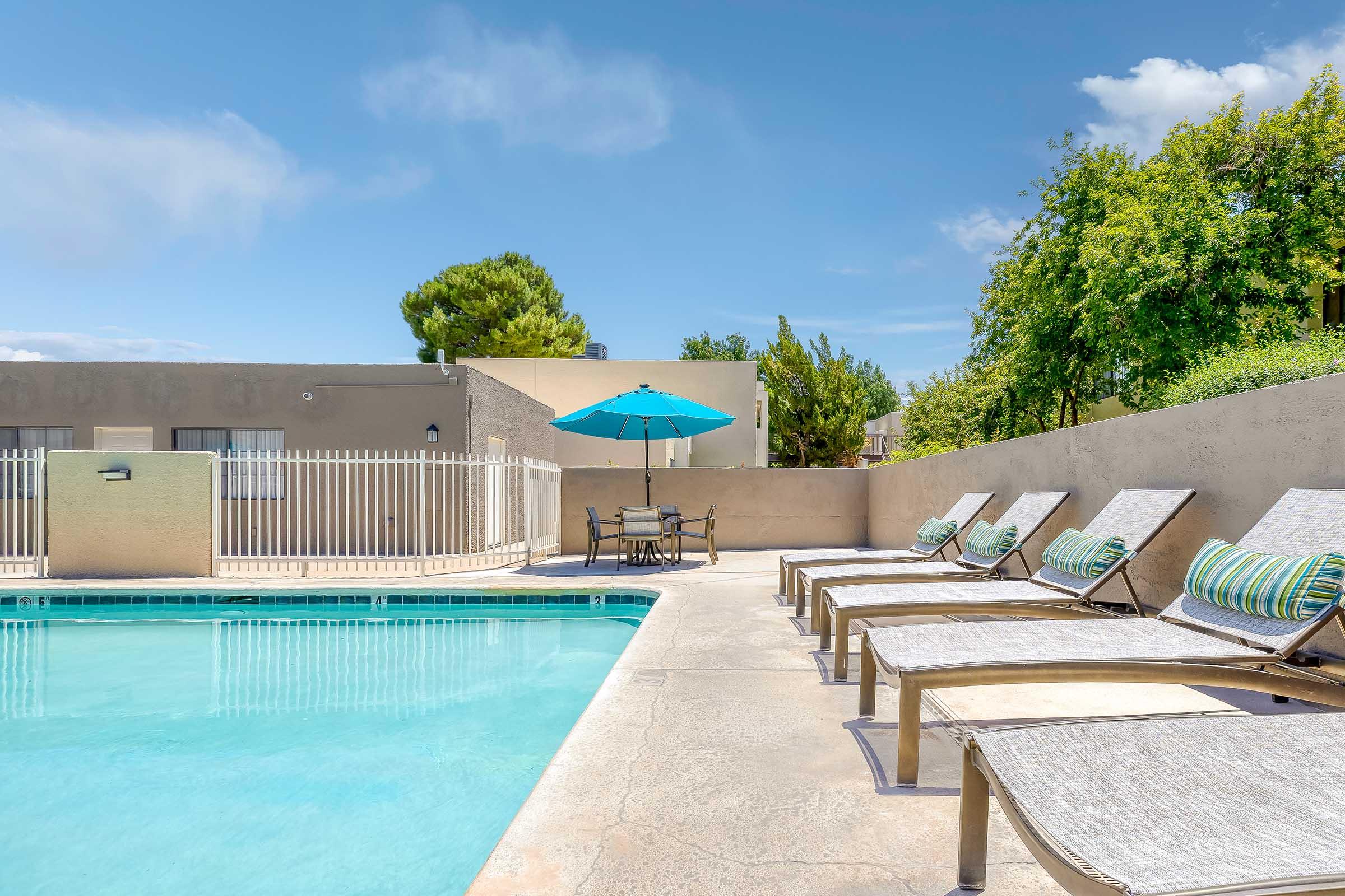 A serene pool area featuring a clear blue swimming pool, surrounded by lounge chairs with patterned cushions. An umbrella provides shade over a small table and chairs, set against a backdrop of lush green trees and a bright sky. The area is enclosed by a white fence, enhancing the tranquil atmosphere.