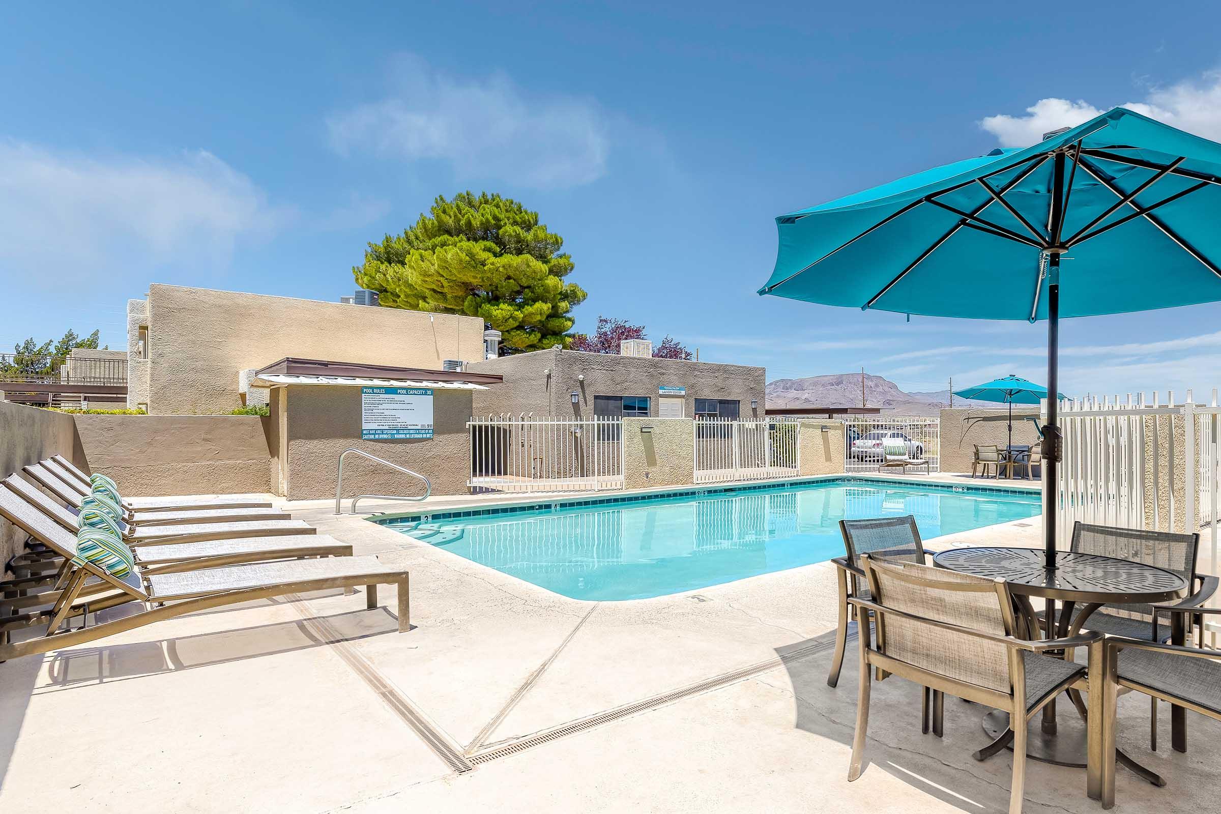 A spacious outdoor pool area featuring a clear blue swimming pool surrounded by sun loungers and a dining table shaded by a large teal umbrella. The backdrop includes light-colored buildings and greenery, with a clear blue sky above.