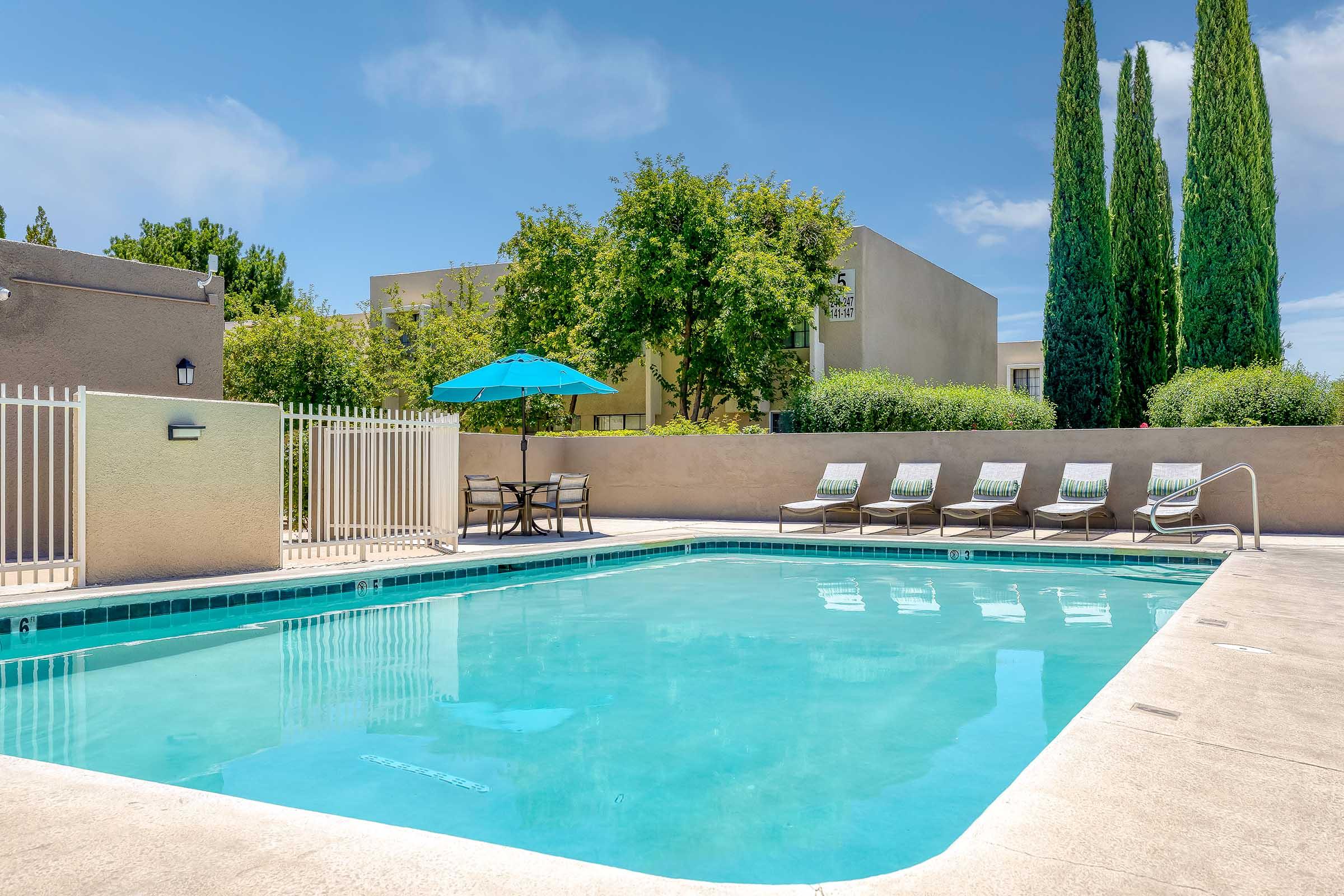 A clear swimming pool surrounded by lounge chairs and a small table with an umbrella. Lush greenery and tall trees are visible in the background, while a light blue sky completes the serene outdoor setting. A fence encircles the pool area.