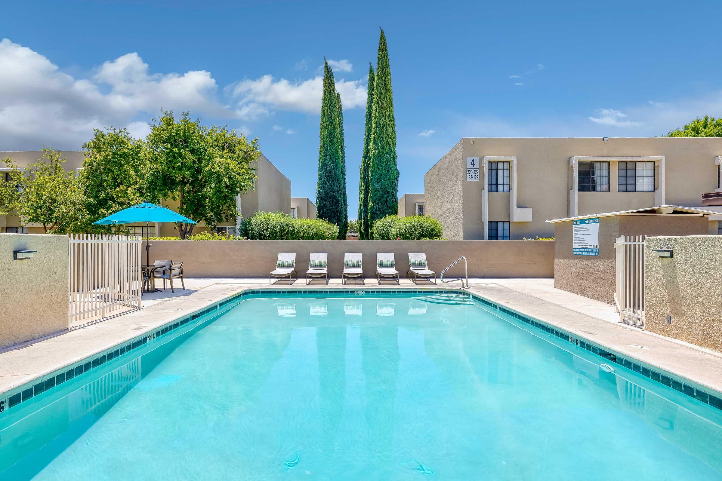 A clear swimming pool surrounded by lounge chairs, with tall cypress trees in the background. The area is enclosed with a beige stucco wall, and there are umbrellas providing shade. The sky is bright and partly cloudy, creating a relaxing outdoor atmosphere.