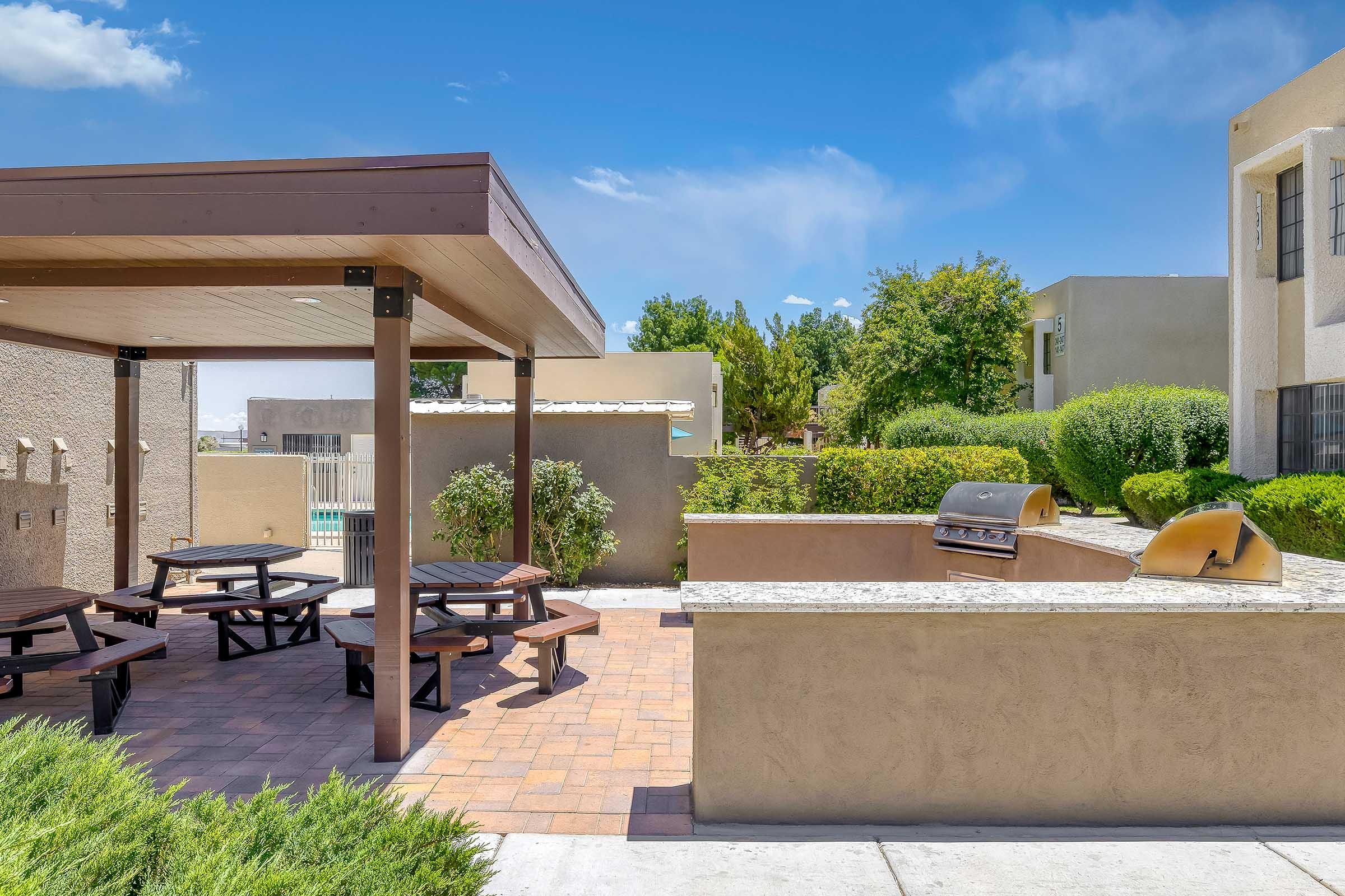 Outdoor communal area featuring a covered patio with picnic tables, a built-in barbecue grill, and lush greenery. A pool is visible in the background, surrounded by residential buildings under a clear blue sky.