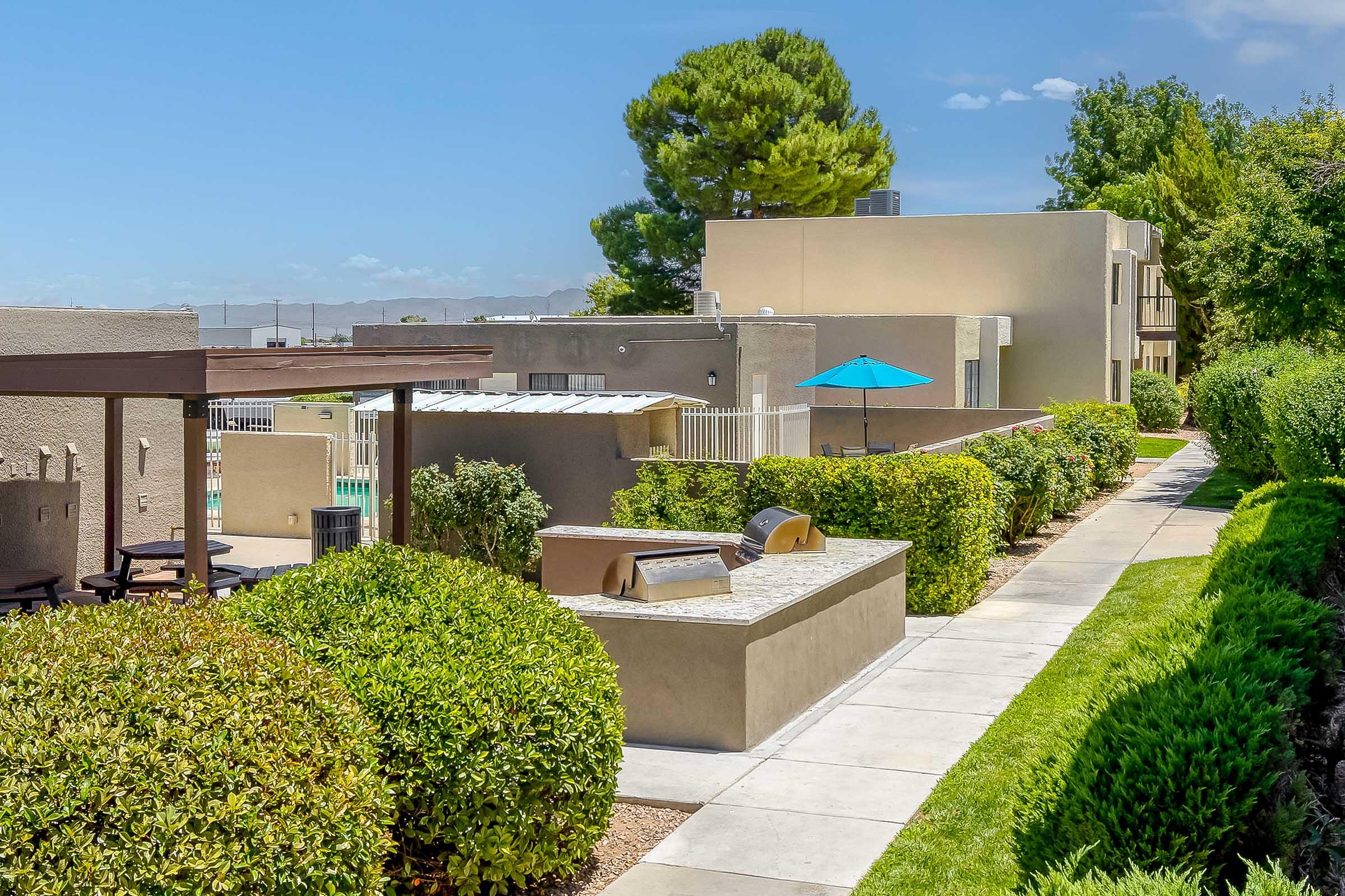 Well-maintained pathway lined with green hedges, leading to modern apartment buildings. In the foreground, a grill area with seating and a blue umbrella beside a pool area is visible, all under a clear blue sky. The scene conveys a relaxing outdoor living space.