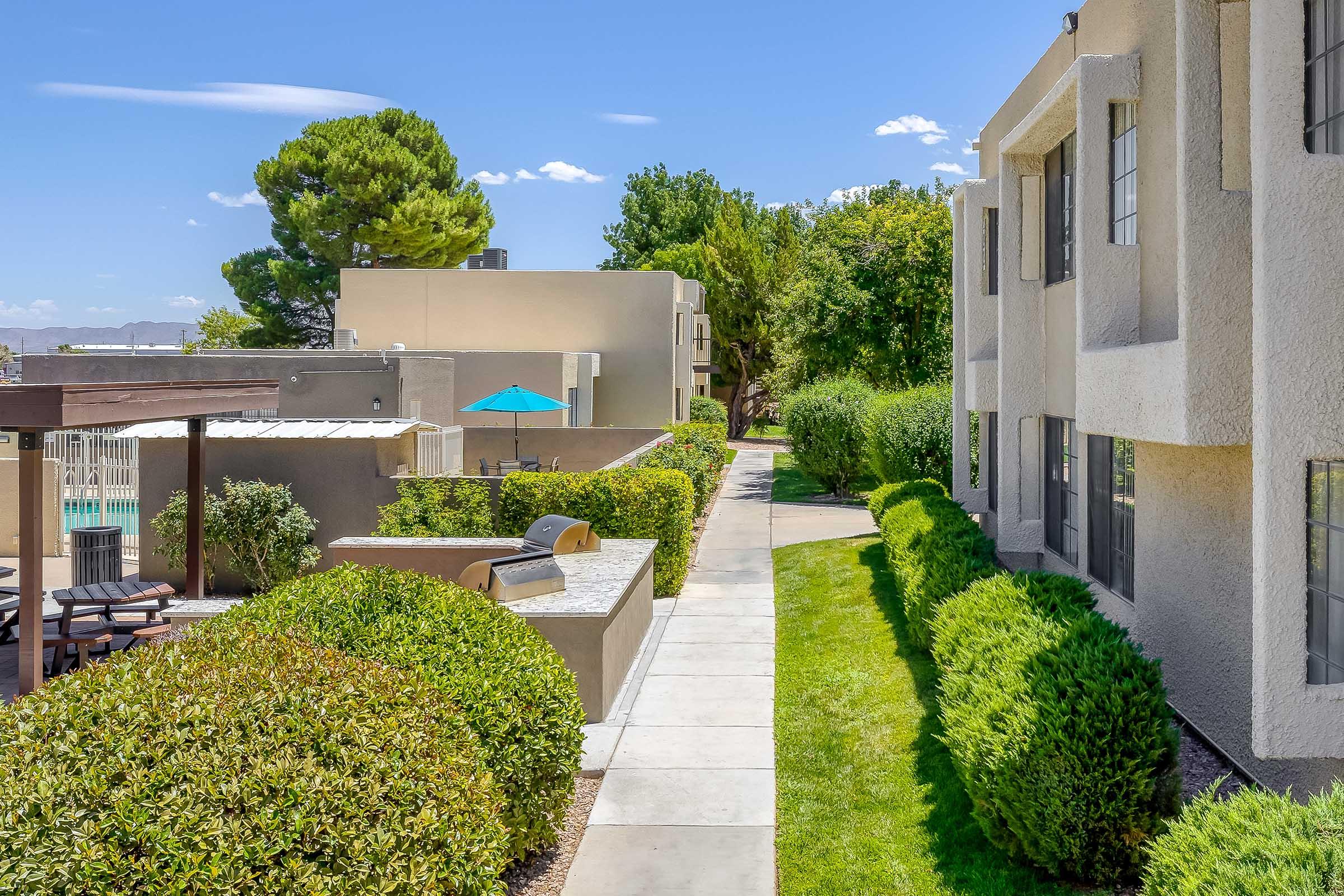 A well-maintained outdoor pathway lined with green hedges, leading to beige apartment buildings. In the background, a pool area with a shaded seating space and an umbrella is visible, surrounded by trees under a clear blue sky.