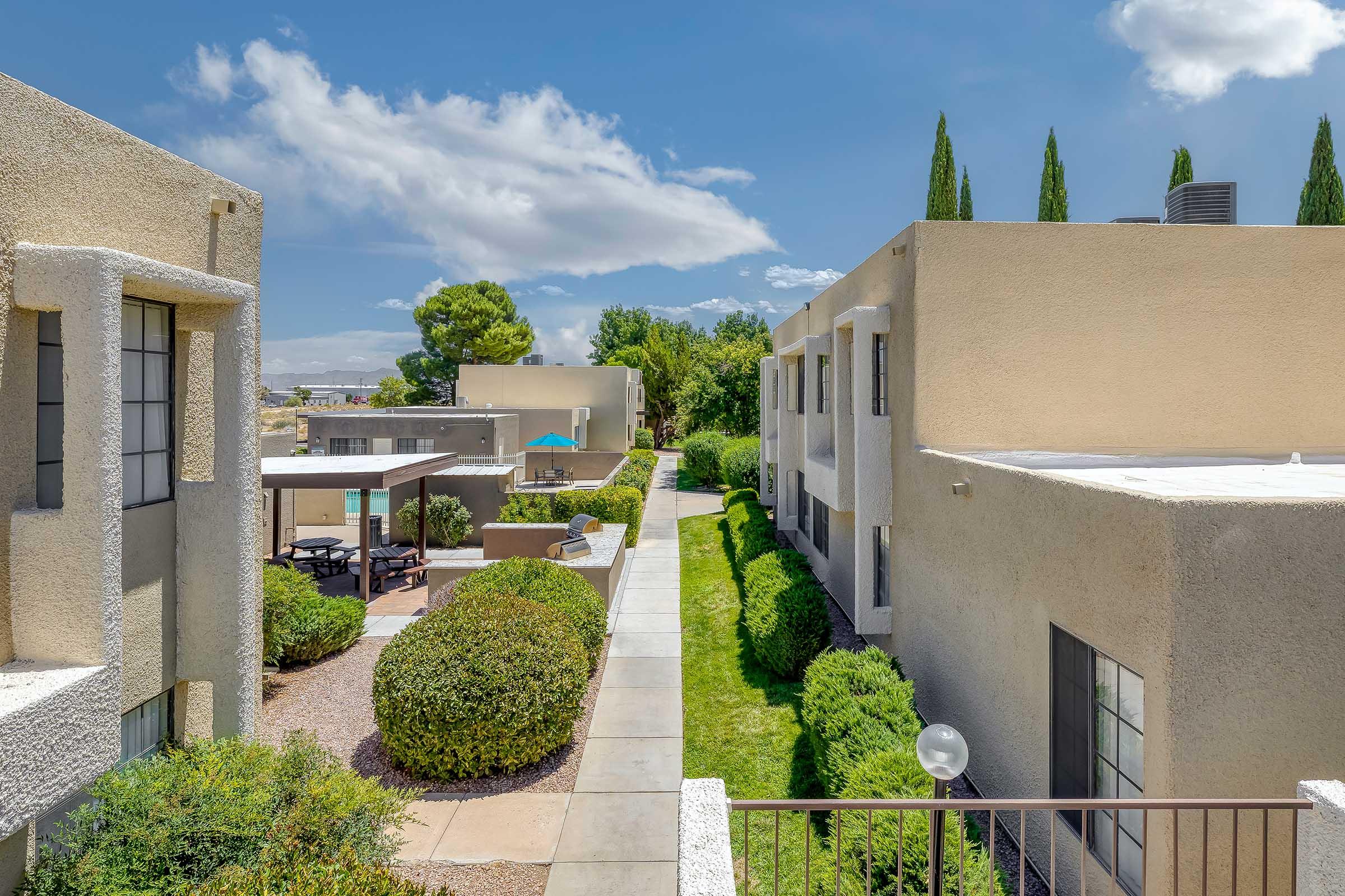 A peaceful courtyard scene featuring landscaped greenery and walking paths between two buildings. The left side shows seating areas and a swimming pool in the background. Bright blue sky with scattered clouds enhances the serene atmosphere.