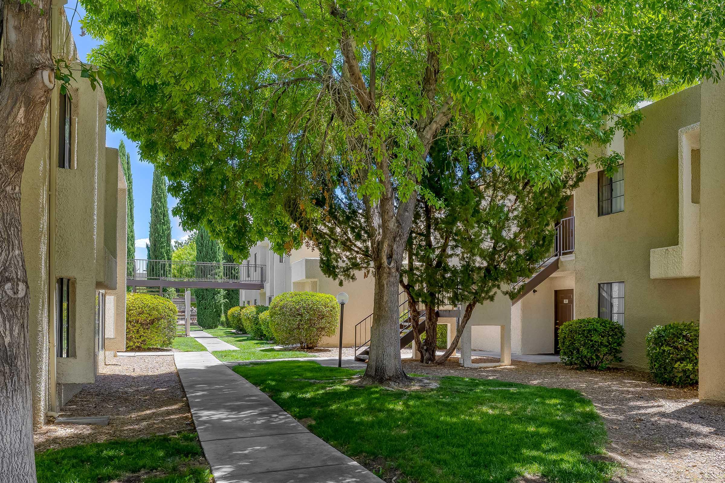 Lush green landscaping surrounds a pathway leading through an apartment complex. The scene features well-maintained buildings with balconies, vibrant trees providing shade, and neatly trimmed bushes under a clear blue sky. A bridge connects two sections of the complex in the background.