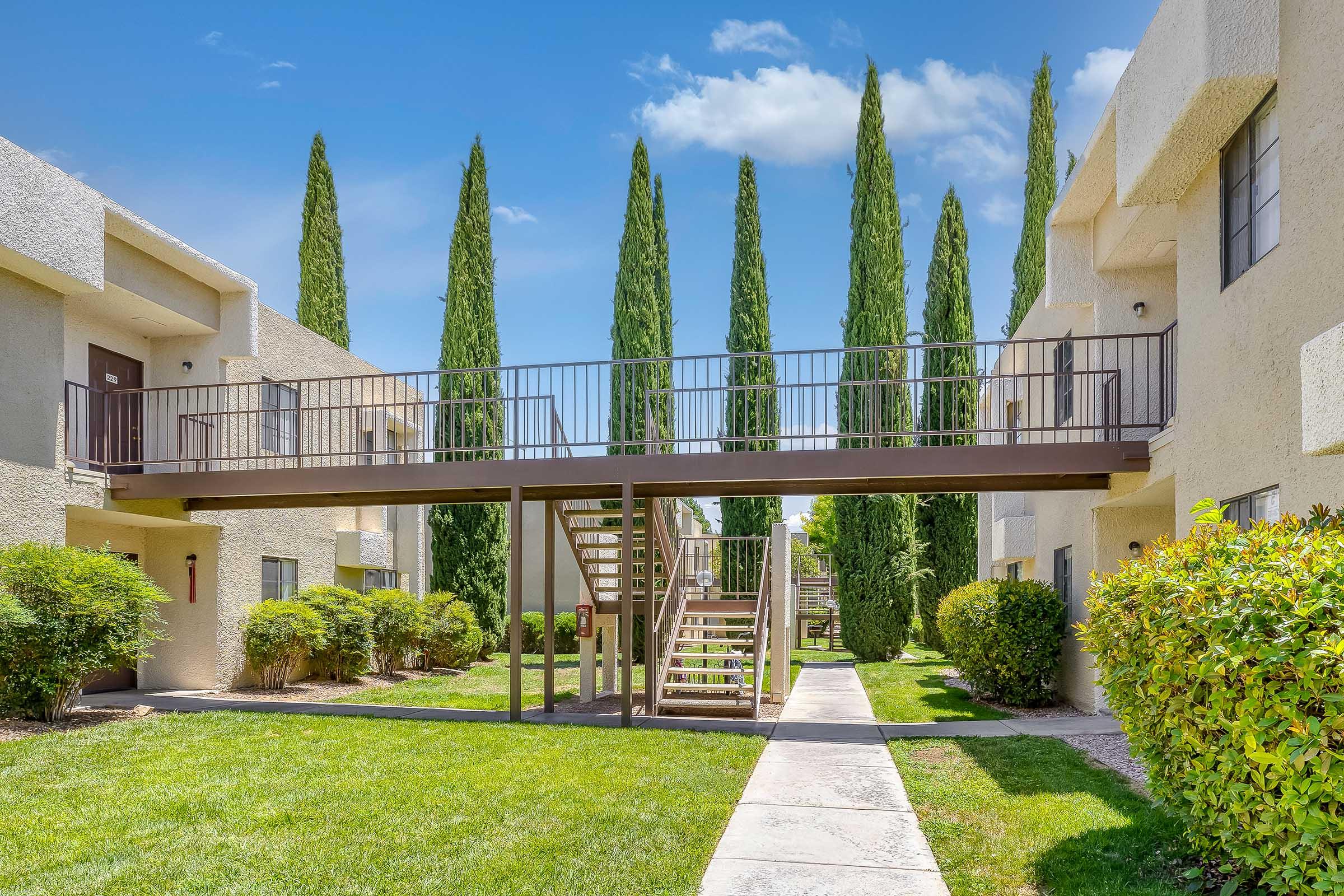 A view of an apartment complex featuring two-story buildings with light-colored walls. There is a raised wooden walkway connecting the buildings, surrounded by neatly trimmed green shrubs and tall, slender cypress trees. The sky is bright blue with a few clouds, creating a cheerful atmosphere.