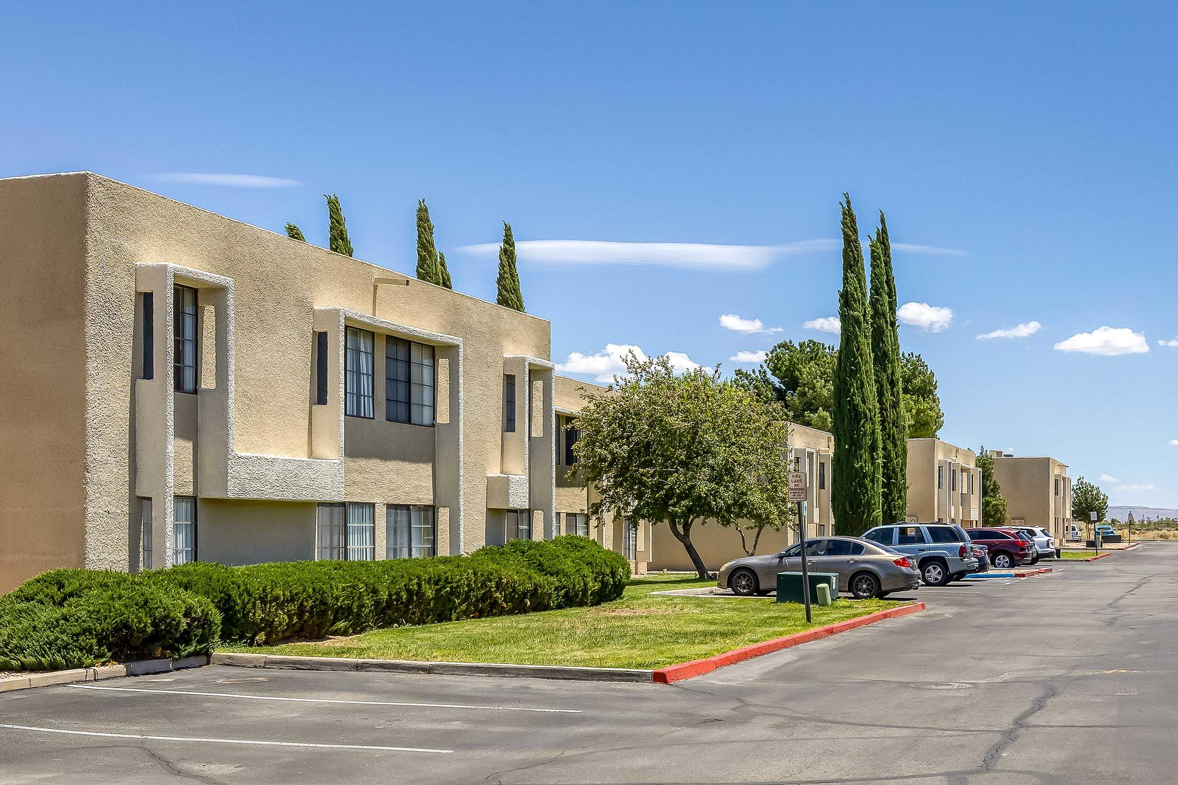 A view of a residential area featuring beige-colored apartment buildings alongside a paved parking lot. Neatly trimmed bushes and trees are present, with several cars parked. The sky is bright blue with some clouds, creating a pleasant atmosphere.