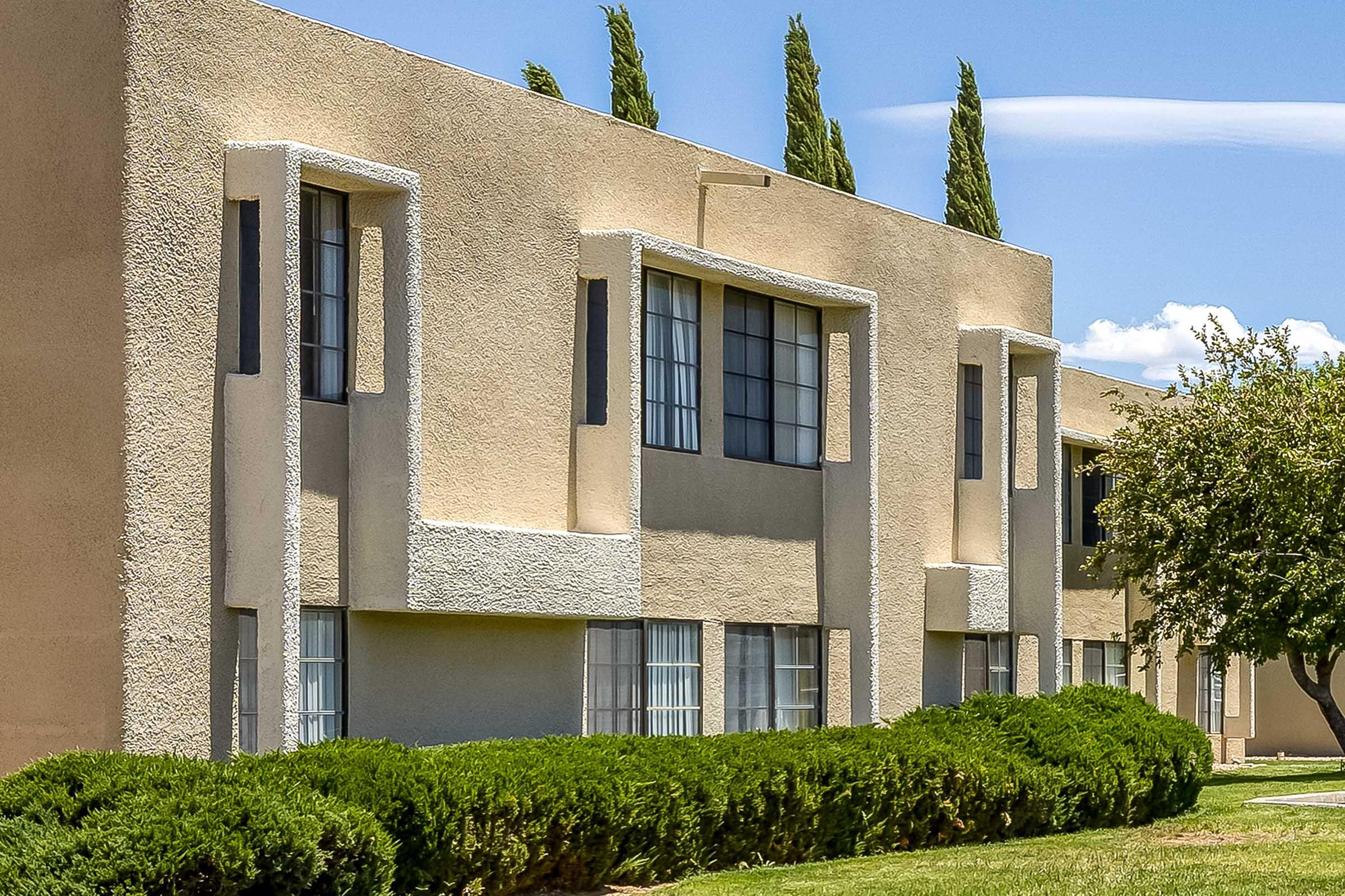 A modern apartment building featuring a beige exterior with white accents. The structure has large windows and is surrounded by well-maintained greenery, including neatly trimmed bushes and trees. The sky is partly cloudy, adding a contrasting blue backdrop.