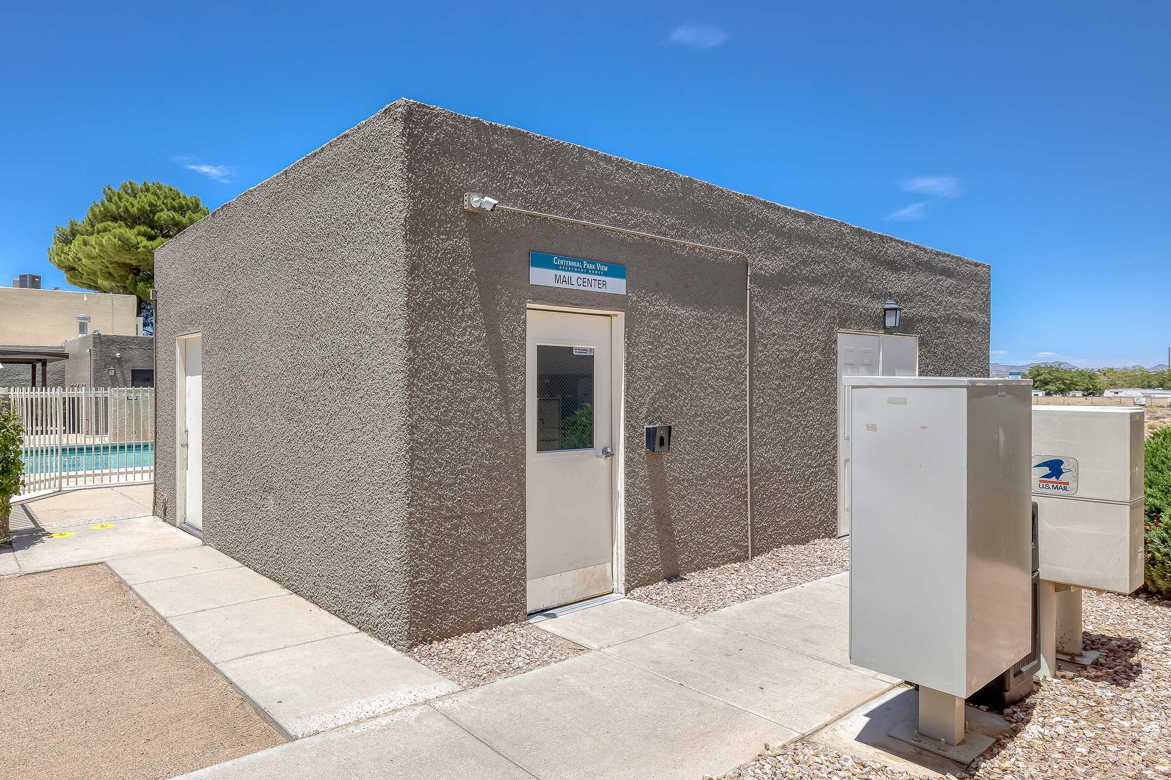 A small gray building labeled "Mail Center" with two doors, surrounded by gravel, near a swimming pool. There are utility boxes next to the building, and landscaping features like shrubs and trees are visible in the background, under a clear blue sky.