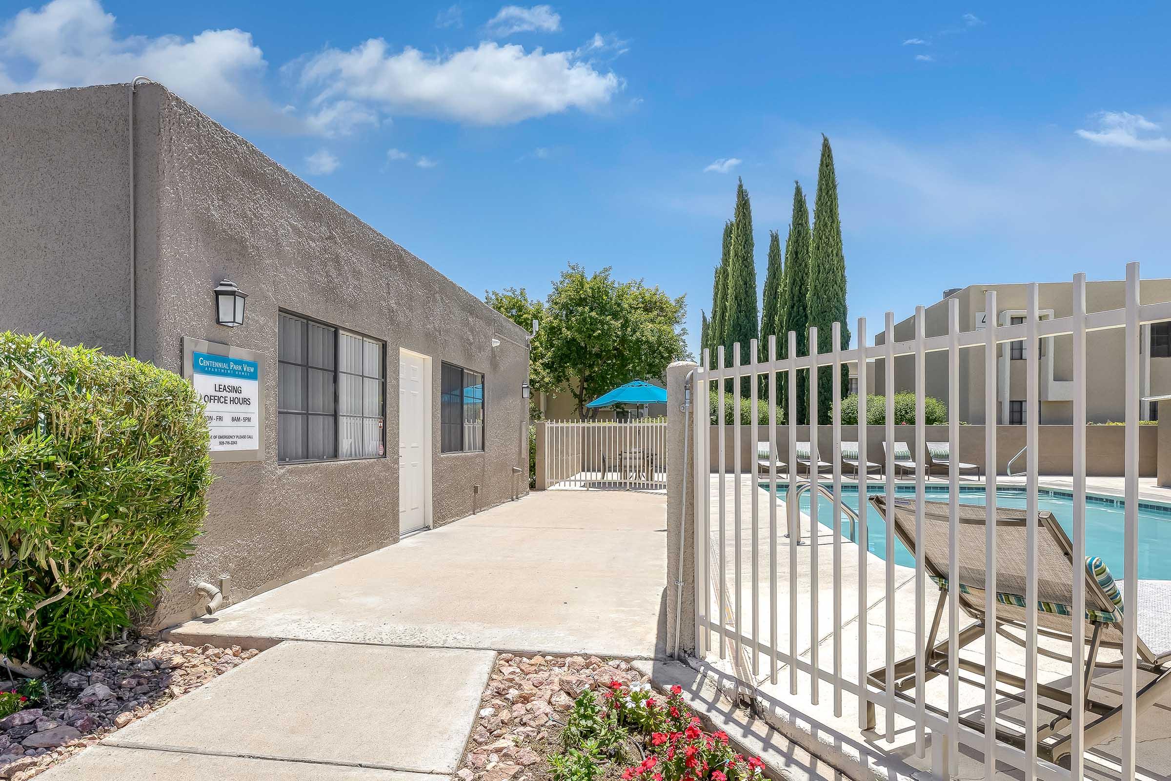A view of a swimming pool area at an apartment complex, featuring a fenced-off pool with lounge chairs. Nearby, there's a sign indicating the leasing office and tall cypress trees lining the background under a clear blue sky. Flowerbeds with colorful plants are seen along the pathway.