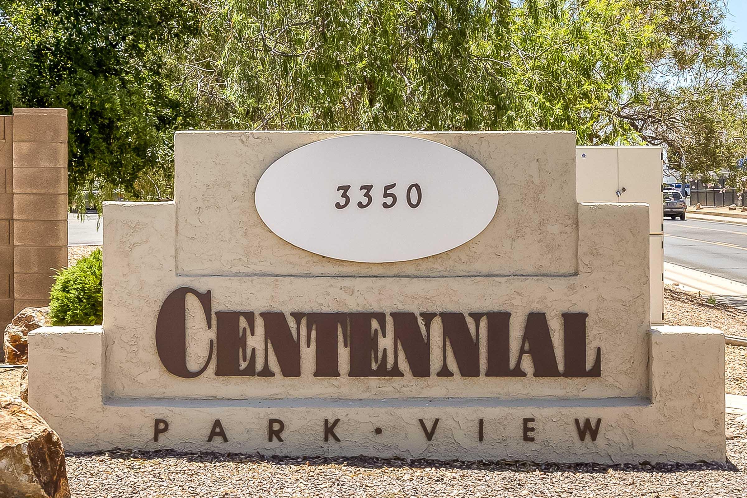 Sign for "Centennial Park View" with the address "3350" prominently displayed. The sign features a beige background with brown lettering, surrounded by landscaping and a clear view of the street in the background.