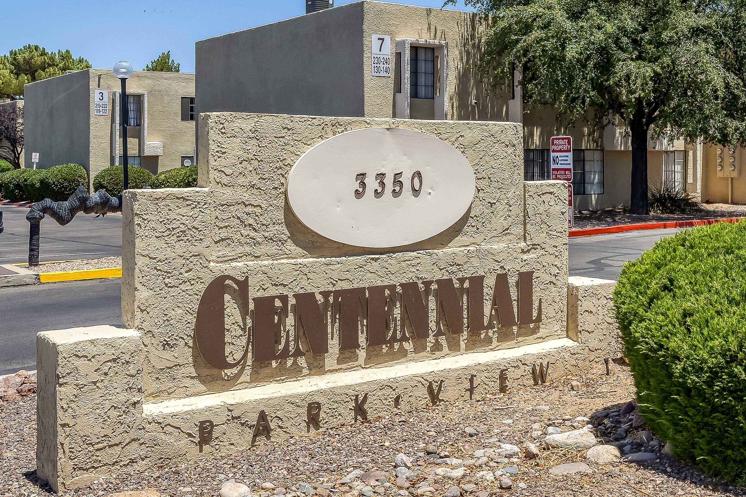 A stone sign reads "CENTENNIAL PARK VIEW" with the address "3550" prominently displayed. The sign is surrounded by green shrubs and landscaping, with buildings in the background. Clear blue sky visible overhead.