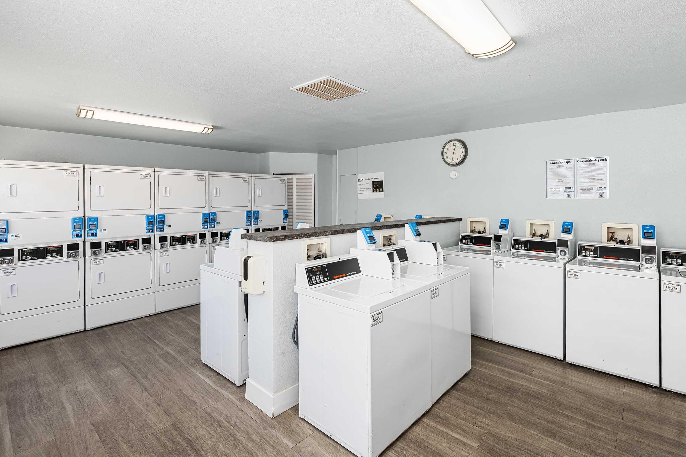 A clean and well-lit laundry room featuring several white washing machines and dryers arranged neatly against the walls. There are informational posters on the wall and a clock mounted above. The floor has a modern wood-like finish, creating a welcoming atmosphere for residents.