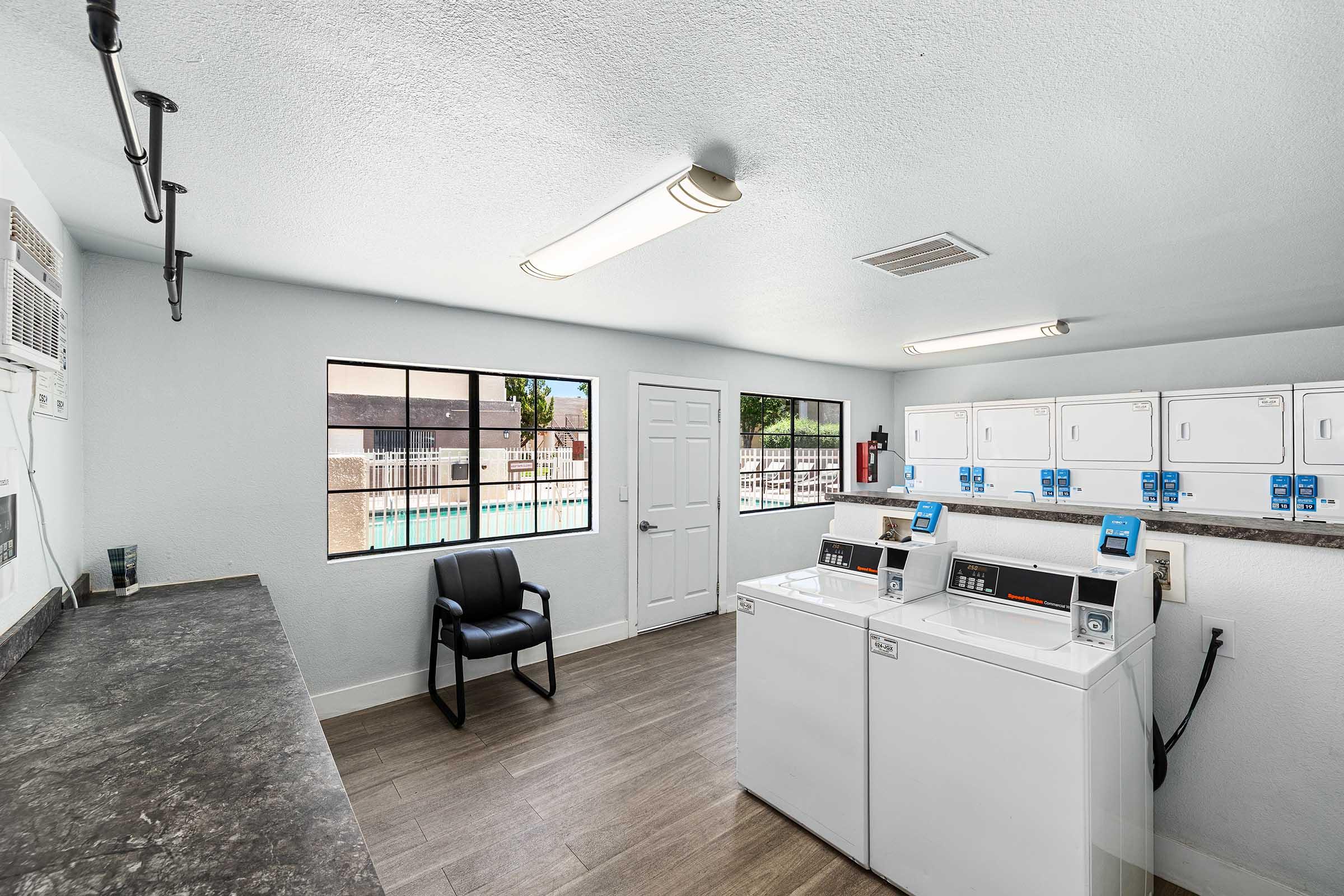 A clean and bright laundry room featuring two white washing machines, a countertop, a chair, and large windows. The decor is simple with gray walls and wooden flooring, and there's a door leading outside, providing a view of the pool area.