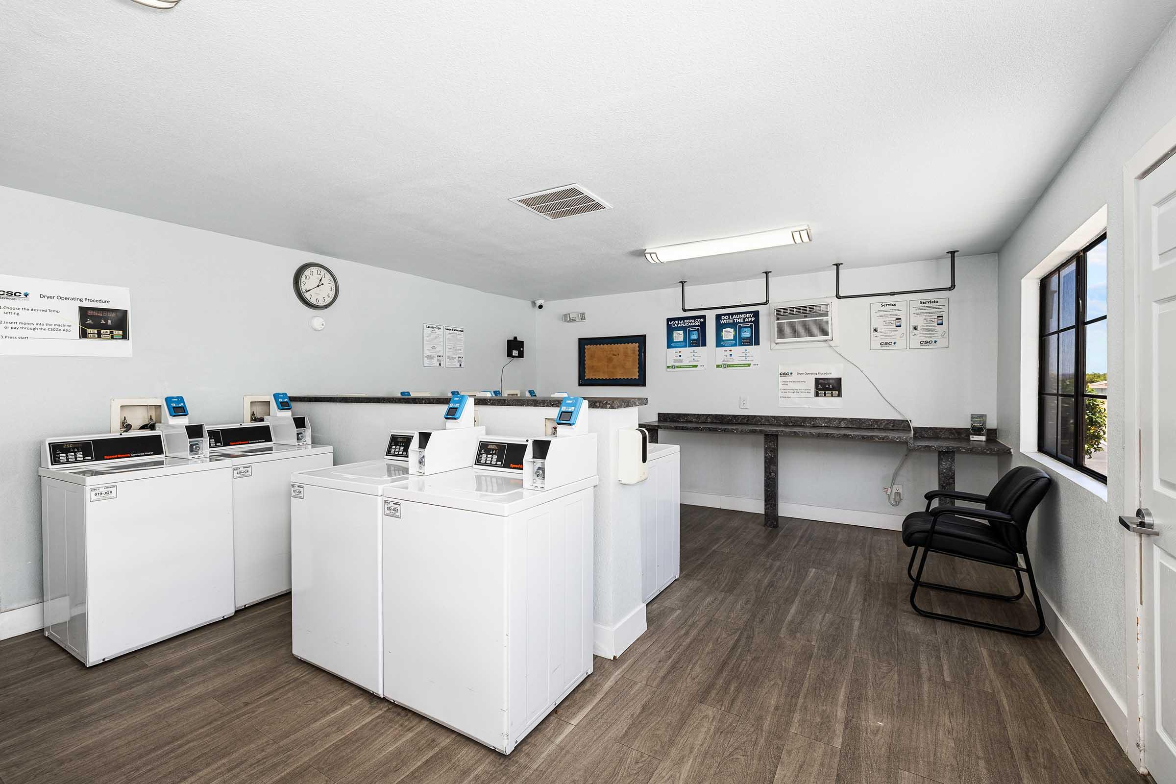 A clean and well-lit laundry room featuring three washing machines, a wall clock, and a countertop. There's a chair against the wall and informational posters visible. The atmosphere is tidy and organized, with windows allowing natural light to enter.