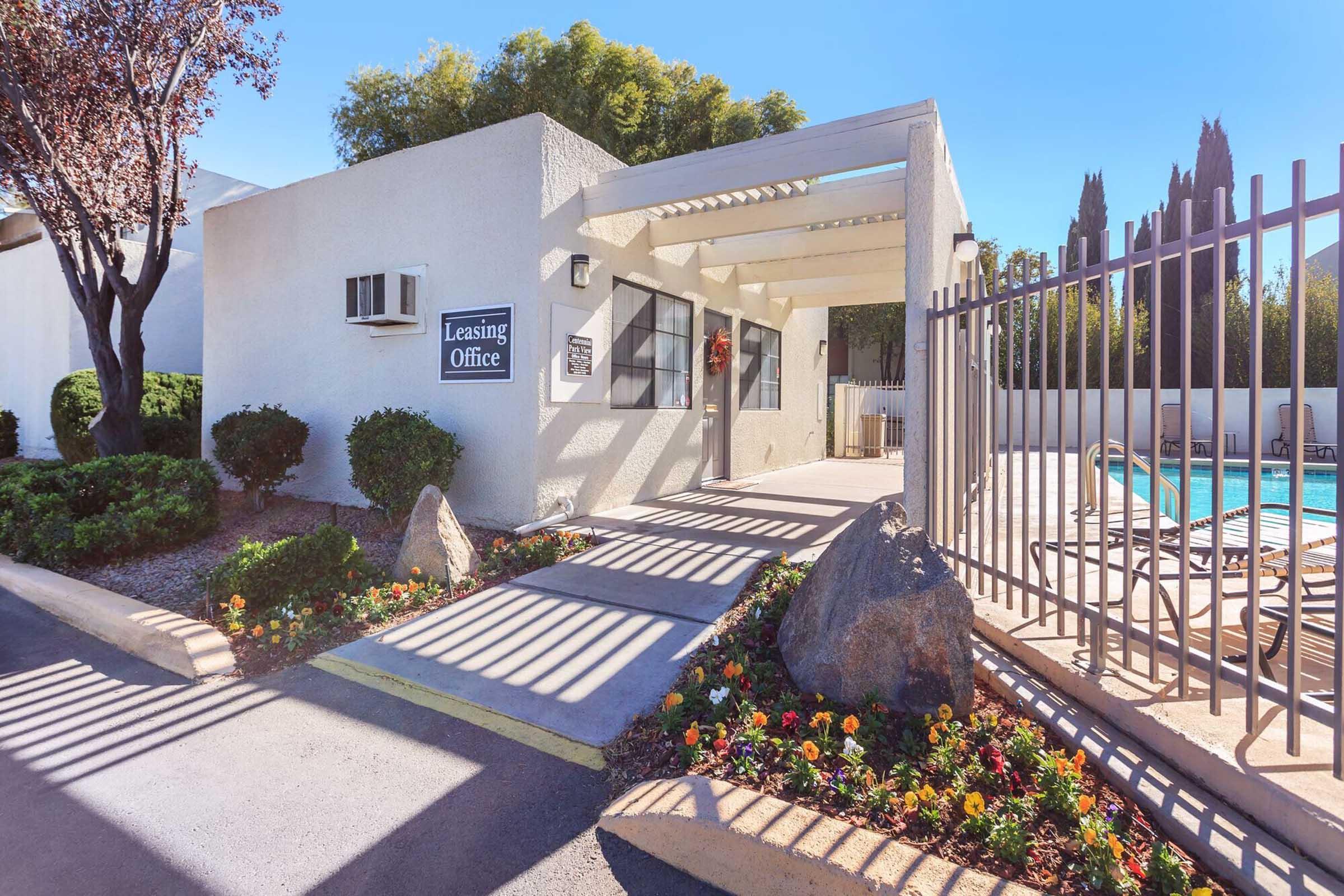 A leasing office with a covered entrance, surrounded by manicured landscaping and flowers. A pool area is visible in the background, with a fence enclosing the space under a clear blue sky. Shadow patterns from the structure create an interesting visual effect on the pavement.