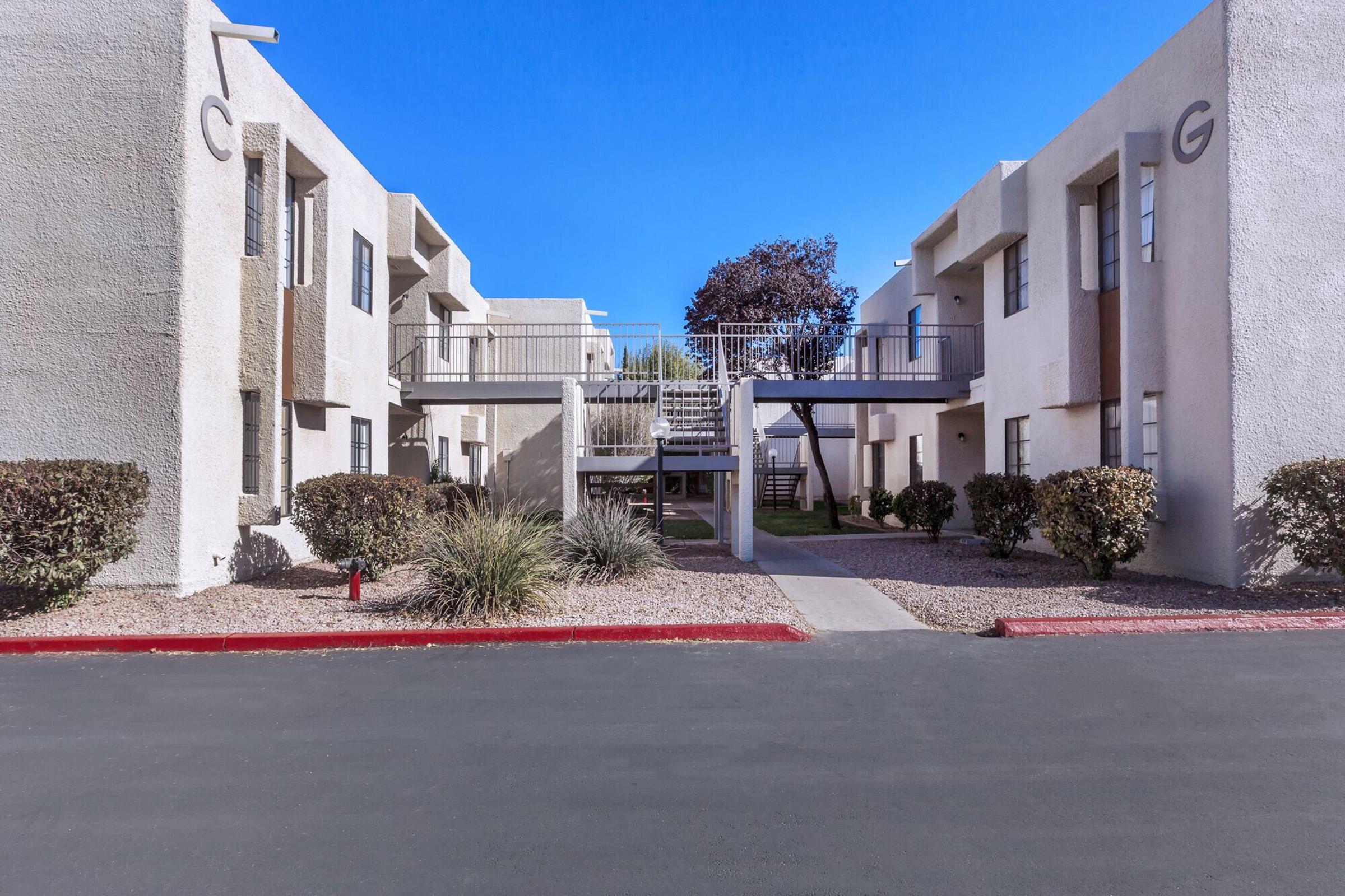 A view of a modern apartment complex featuring two white buildings with balconies and staircases. The walkway is flanked by landscaped shrubs and there is a clear blue sky overhead. The area is paved, indicating a well-maintained outdoor space.