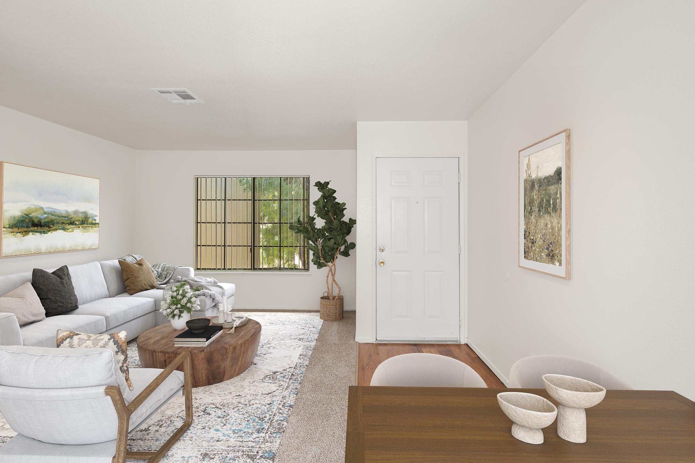 A bright, modern living room featuring a cozy white sectional sofa with decorative pillows, a wooden coffee table, and a large window letting in natural light. The space includes a small dining area with a wooden table and bowls, and decorative art pieces on the walls, enhancing the inviting atmosphere.