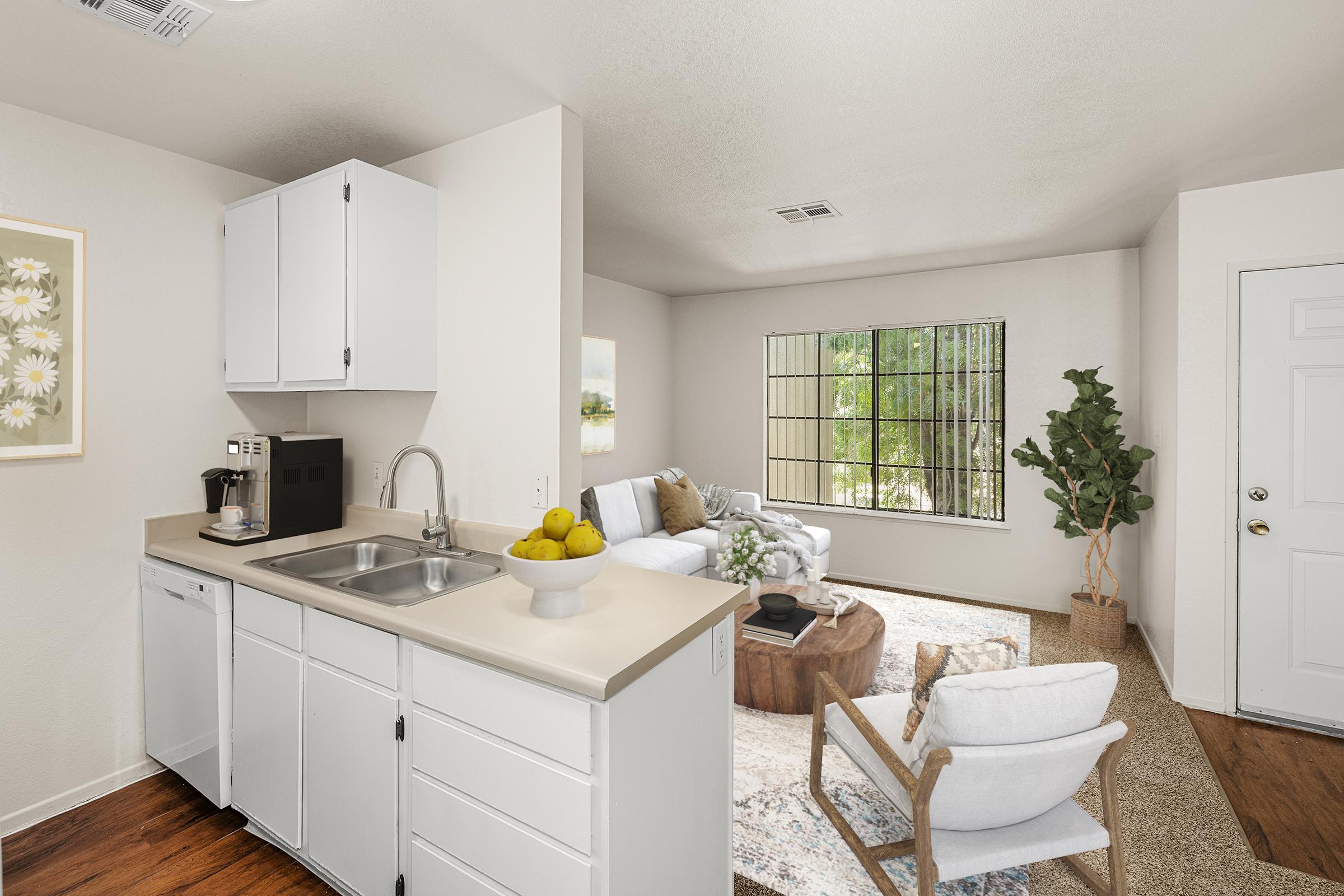 A bright and modern living space featuring a kitchen area with white cabinets and a sink, a cozy living room with a light-colored sofa, a wooden coffee table, and large windows letting in natural light. A small plant adds a touch of greenery, creating a welcoming atmosphere.