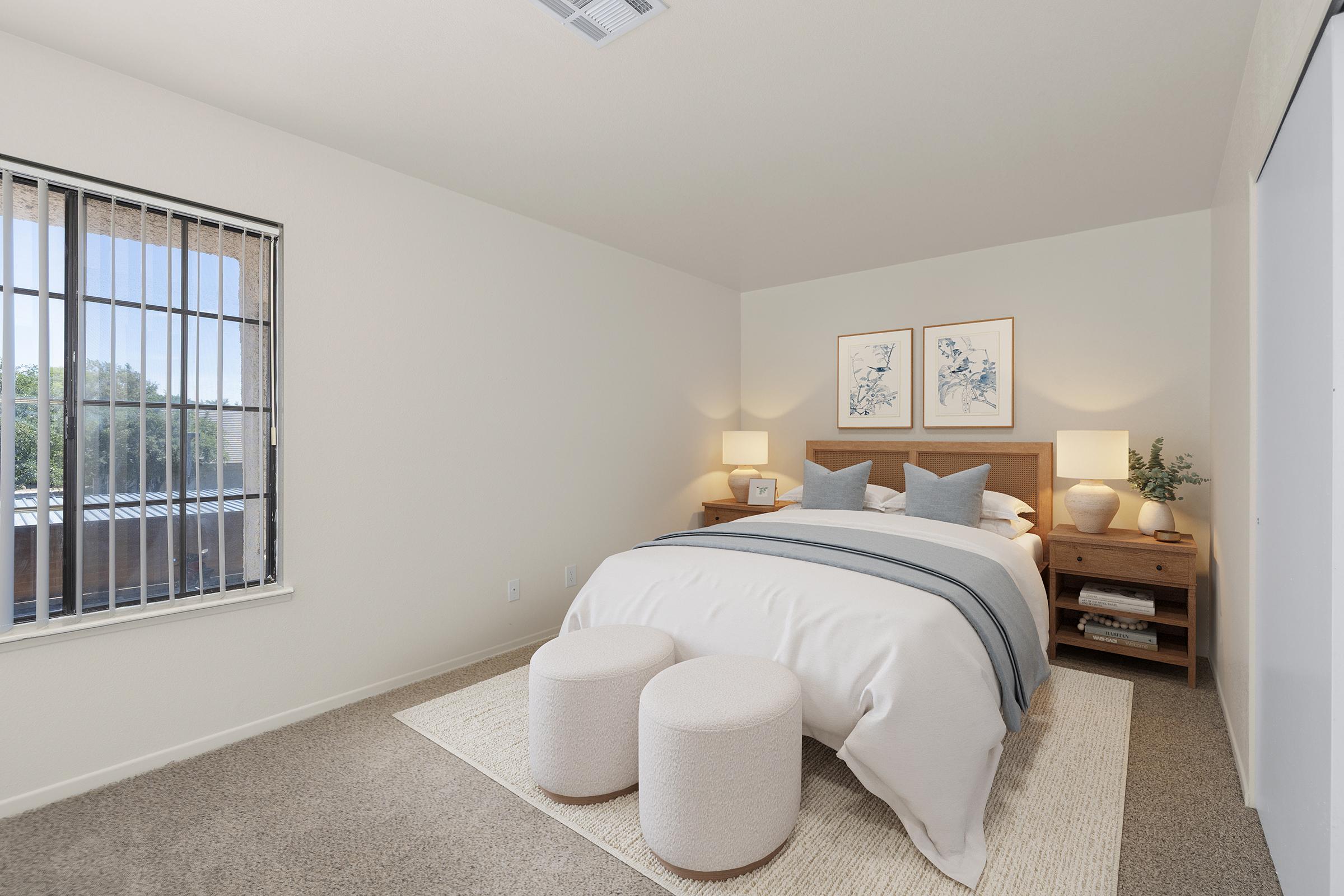 A cozy bedroom featuring a queen-sized bed with soft linens, two bedside tables with lamps, and framed artwork on the wall. The room has a large window with vertical blinds, allowing natural light to illuminate the space, which includes a plush rug and two circular poufs at the foot of the bed.