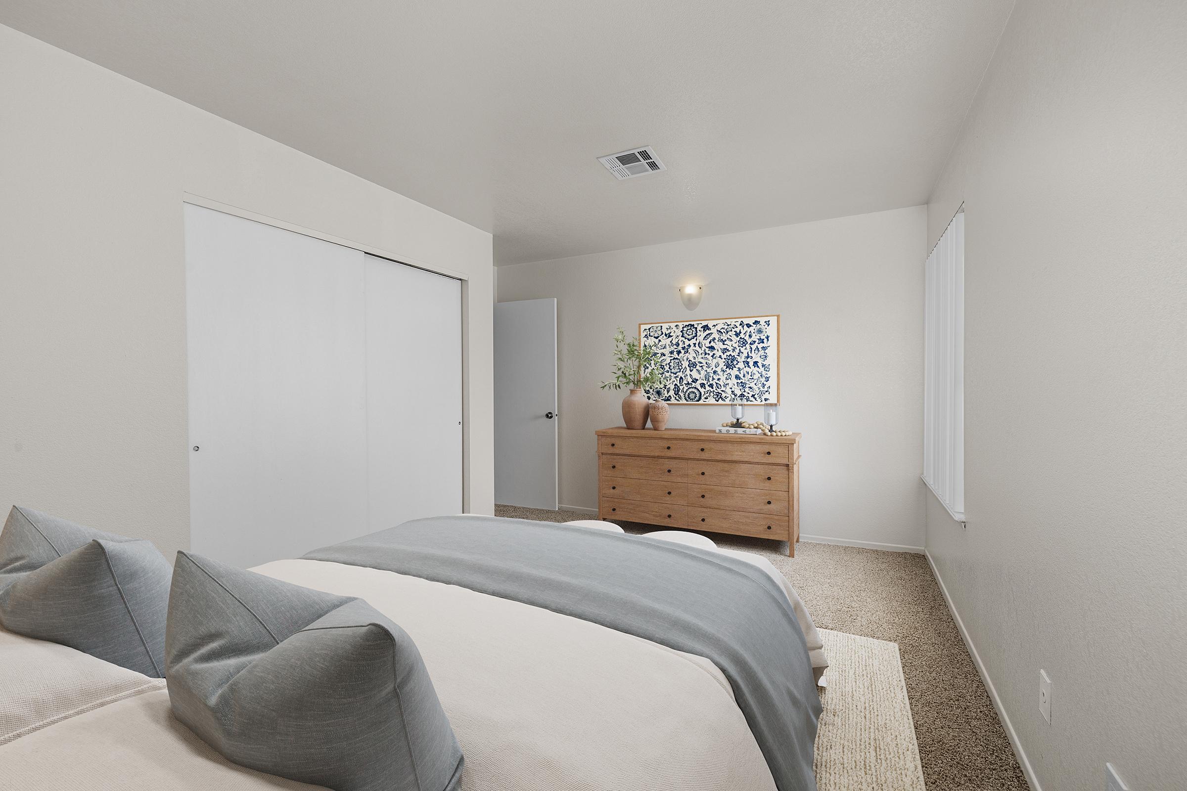 Cozy bedroom featuring a neatly made bed with gray and white bedding, decorative pillows, a wooden dresser with a vase, and a wall-mounted artwork. Natural light filters through a window, while light-colored walls and carpet create a warm atmosphere. A closet door is partially visible on one side.