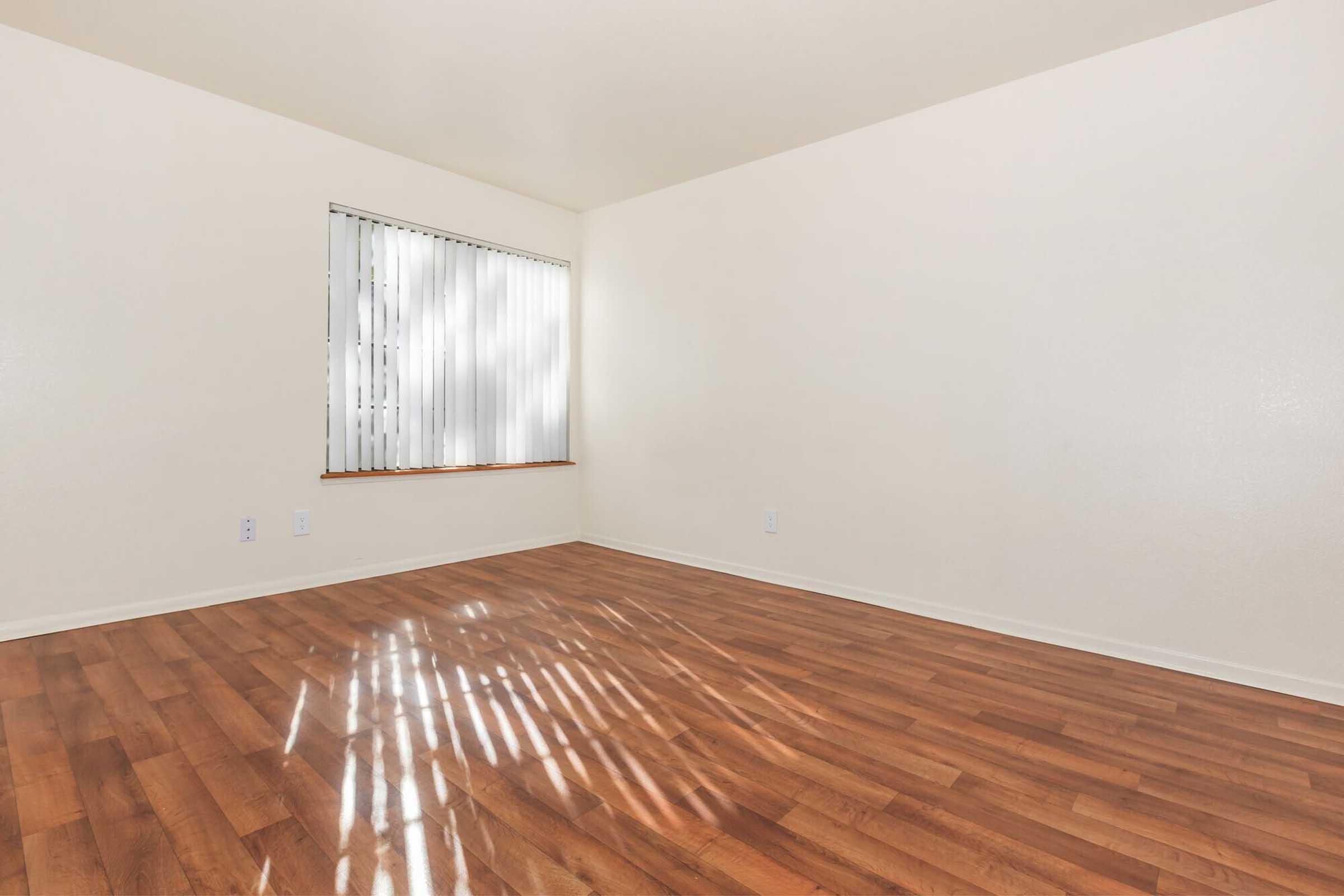 A spacious and empty room featuring light-colored walls and hardwood flooring. A window with vertical blinds allows natural light to enter, creating soft reflections on the floor. The space is clean and ready for furnishing, with electrical outlets on the walls.