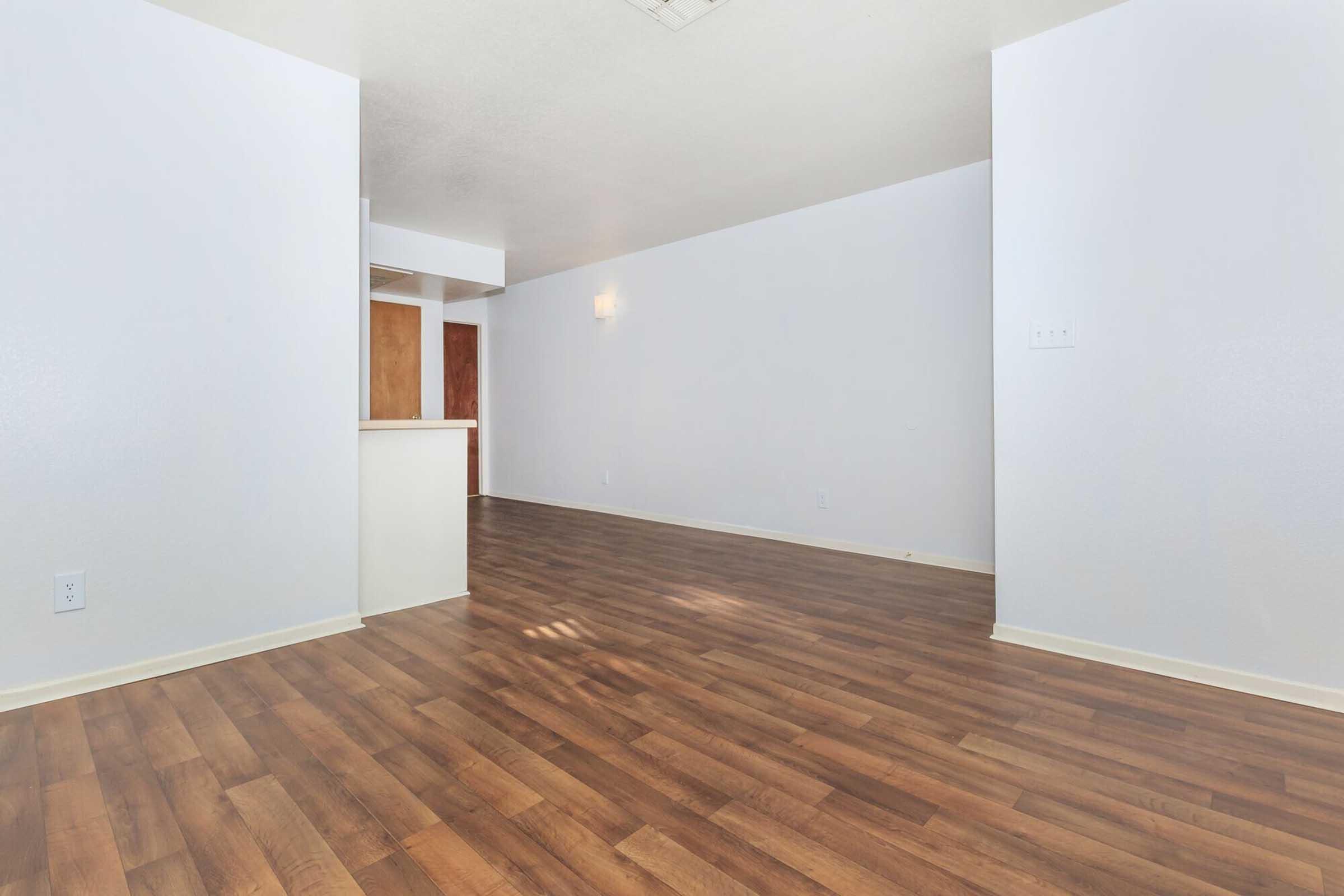 A spacious, empty living area with light-colored walls and a medium brown laminate floor. The room features an open layout with a small kitchen area visible in the background, showcasing minimal furniture and a bright, inviting atmosphere. Natural light enhances the airy feel of the space.