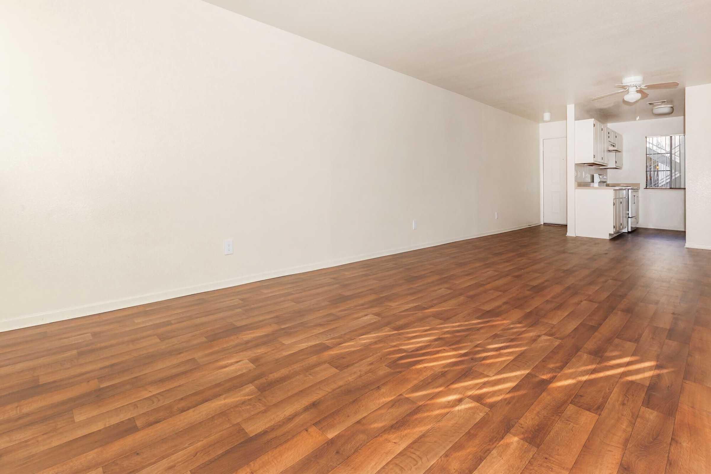 An empty room featuring a wooden floor and light-colored walls. There is a kitchen area in the background with white cabinetry, and a window allowing natural light in. The space is spacious and uncluttered, making it ideal for decoration or furnishing.