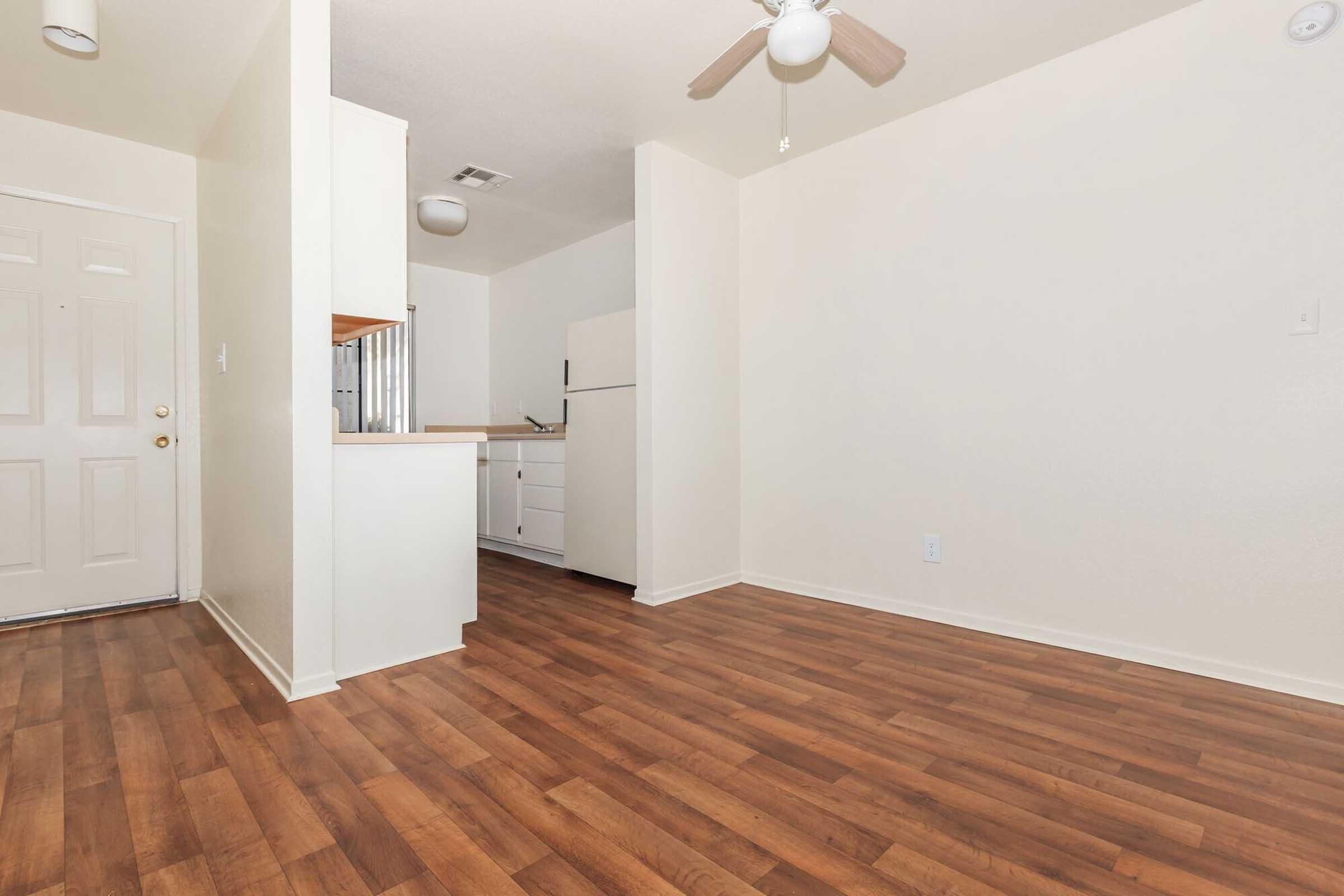 A light-colored room with a wooden floor, featuring a ceiling fan and a door leading outside. To the left, there is a small kitchen area with white cabinets and a refrigerator. The walls and ceilings are painted in soft, neutral tones, creating a spacious and inviting atmosphere.
