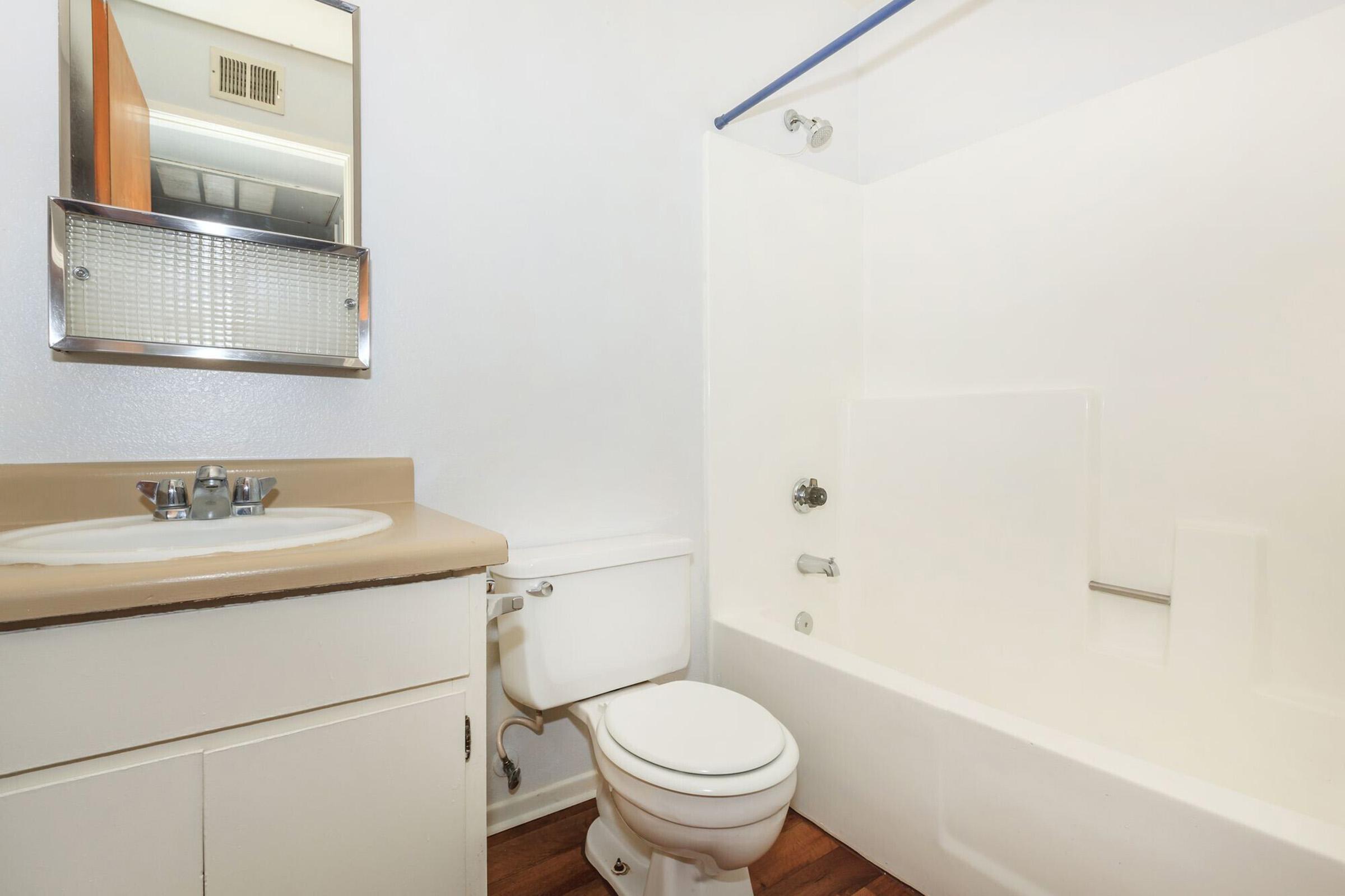 A clean and modern bathroom featuring a white bathtub with a shower, a toilet, and a single sink vanity with a mirror above. The walls are white, and the floor has wood-like flooring. The space is well-lit and tidy, creating a fresh and inviting atmosphere.