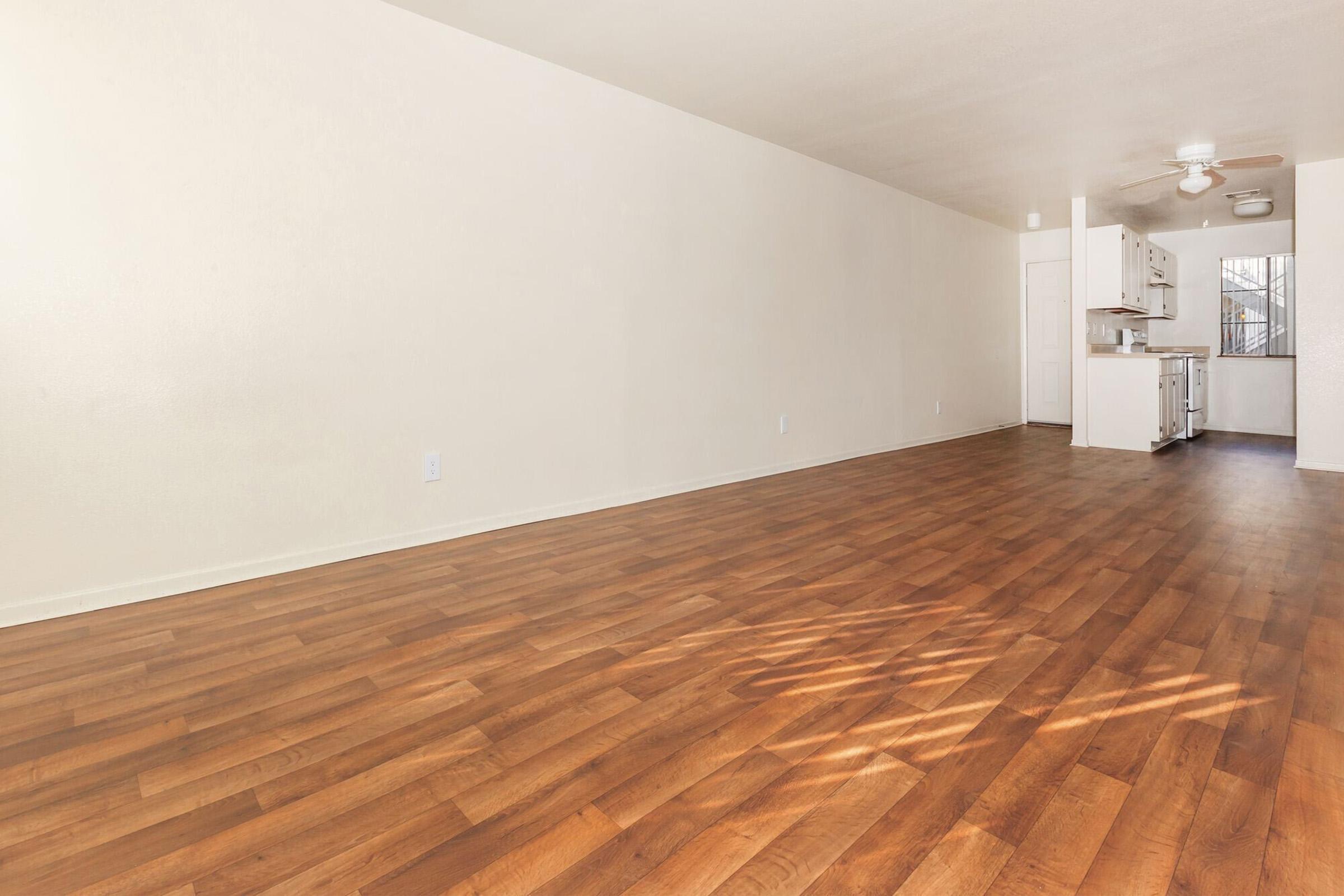 Spacious, well-lit room featuring light-colored walls and a large wooden floor. The area opens to a kitchen with white cabinetry and a window, providing a bright, inviting atmosphere. The layout emphasizes a minimalistic design, suitable for versatile furniture arrangements.