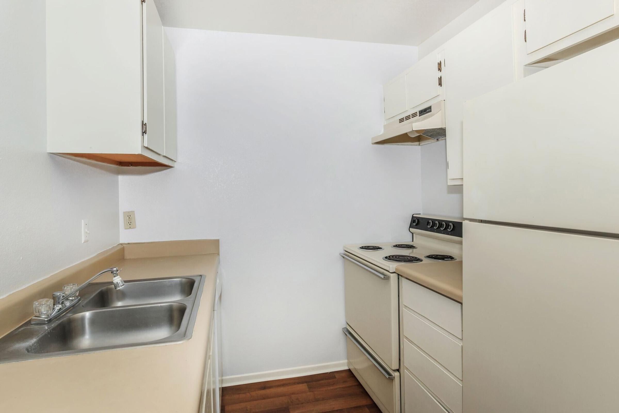 A small, functional kitchen featuring light-colored cabinets, a beige countertop, a double sink, an electric stove with four burners, and a refrigerator. The walls are painted white, and the flooring is a warm-toned wood laminate. The space is organized and designed for efficiency.