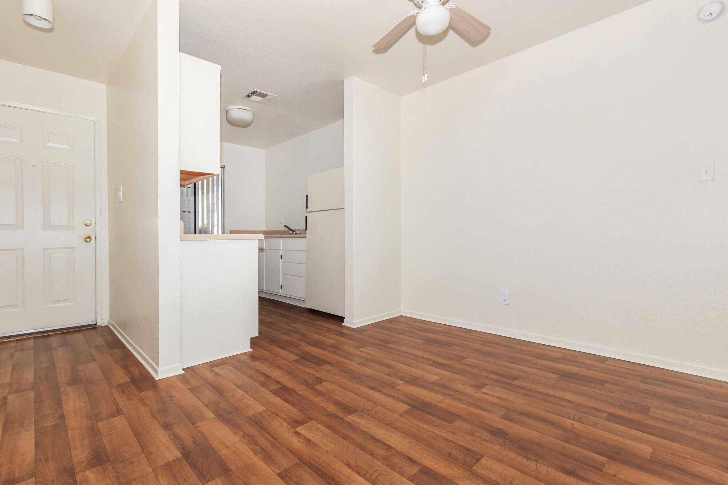 A spacious, empty room with light-colored walls and a wood-like floor. To the left is an entrance door, and on the right, there is a small kitchenette with white cabinets and a white refrigerator. A ceiling fan is visible, contributing to the airy feel of the space.