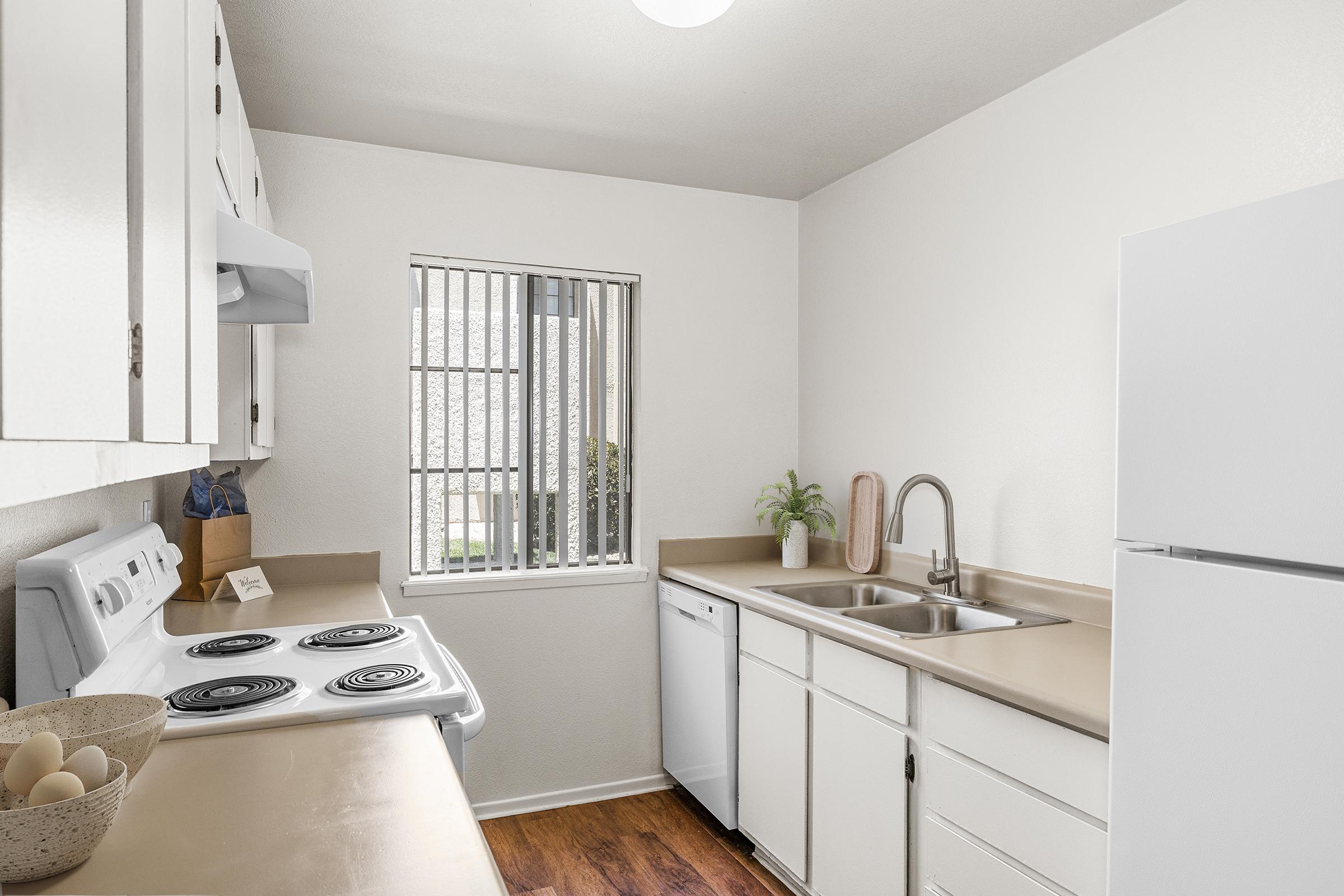 A modern kitchen with white cabinetry, a stove, a refrigerator, and a double sink. The kitchen features a light-colored countertop and wooden flooring. A window with vertical blinds allows natural light to enter, while a small plant and a kitchen accessory are placed on the counter.