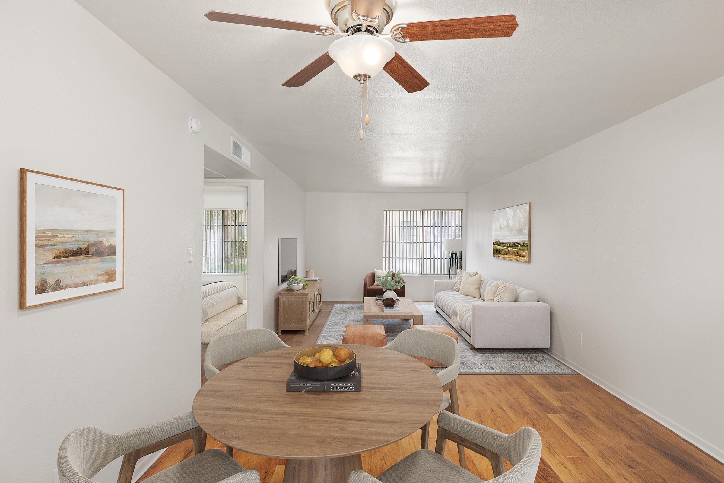 A well-lit, modern living space featuring a round wooden dining table with four chairs, a cozy sofa, and a stylish coffee table. The walls are painted in neutral tones, and there are decorative artworks. Large windows allow natural light to fill the room, creating a welcoming atmosphere.