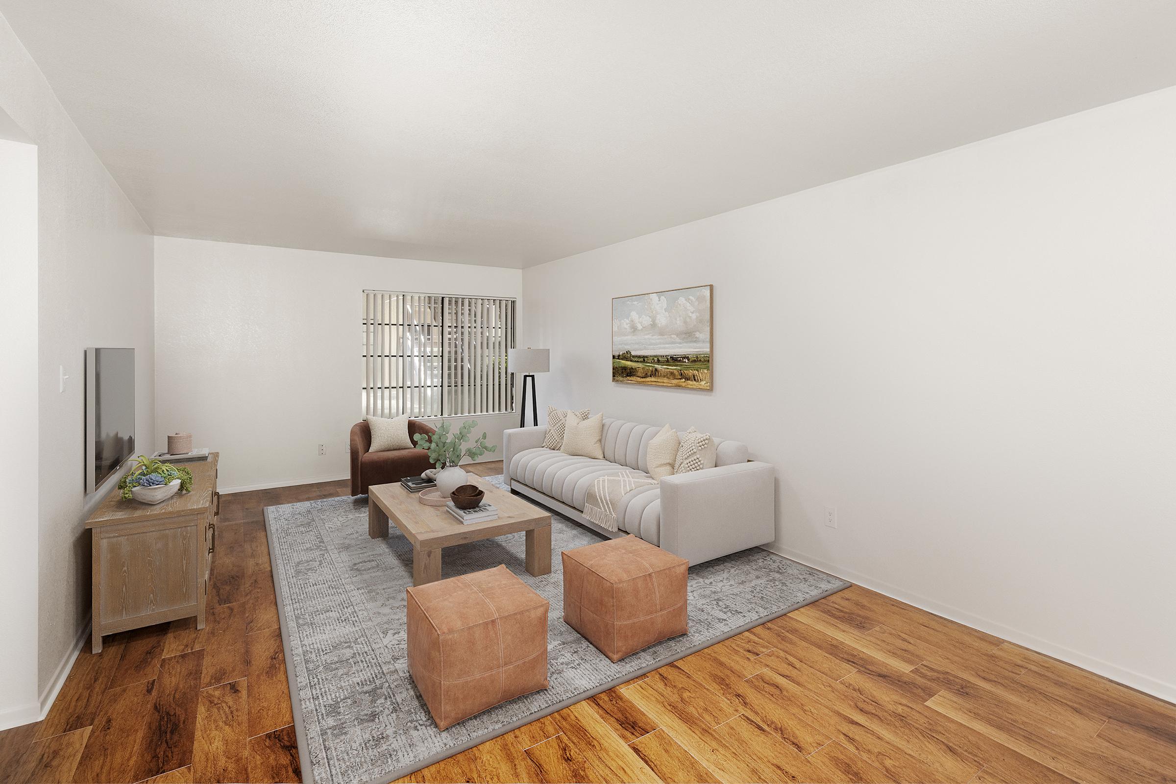 A light-filled living room featuring a comfortable cream sofa, a dark brown armchair, and two orange ottomans. A wooden coffee table sits on a gray area rug, while a TV is mounted on a wall. A painting of a landscape decorates the wall, and a plant is placed on the coffee table.