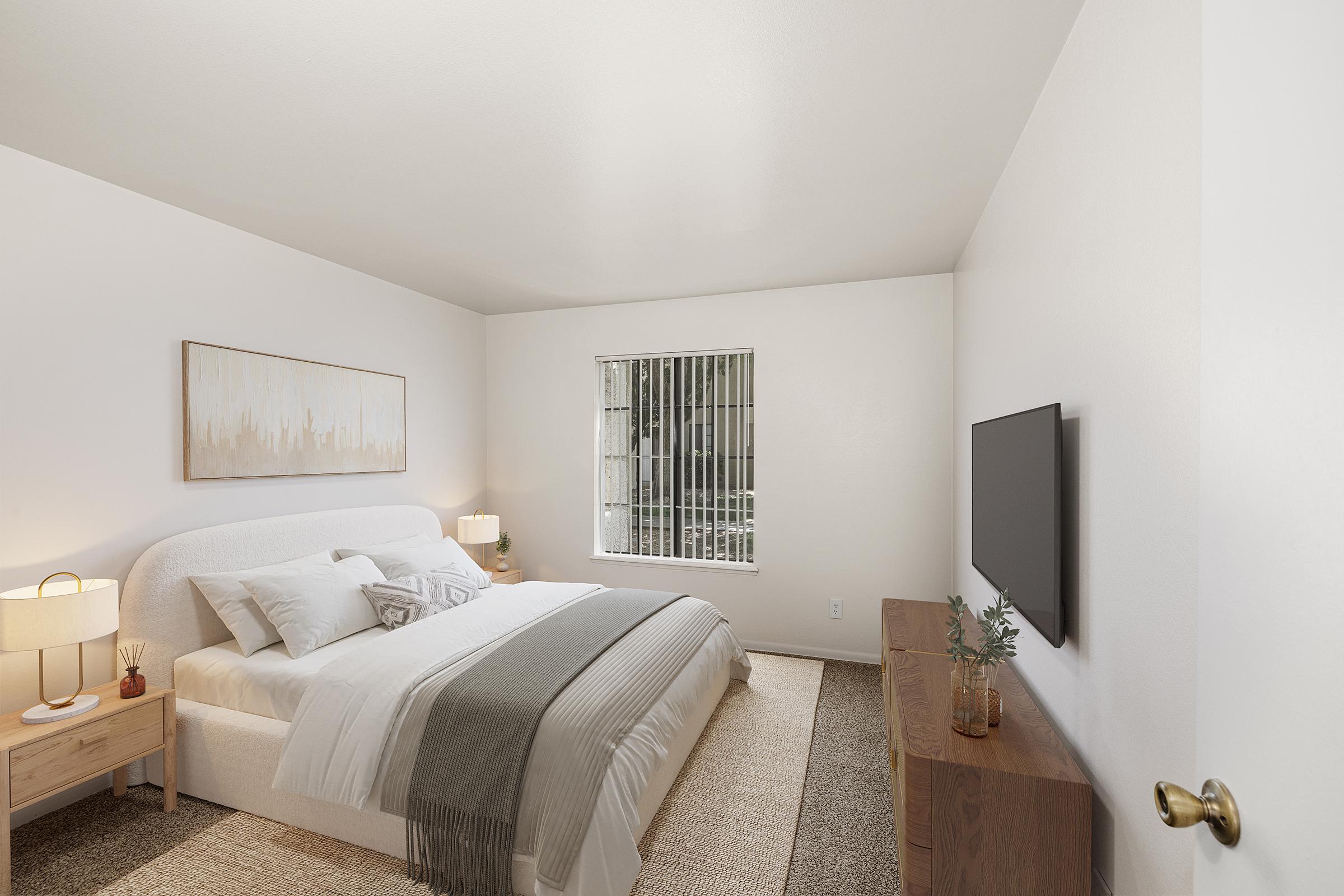 A modern, airy bedroom featuring a large bed with white bedding, a gray throw blanket, and two bedside lamps. There is a wooden dresser with a flat-screen TV mounted on the wall. A window with vertical blinds allows natural light to enter, and the walls are painted in a light color, creating a calming atmosphere.
