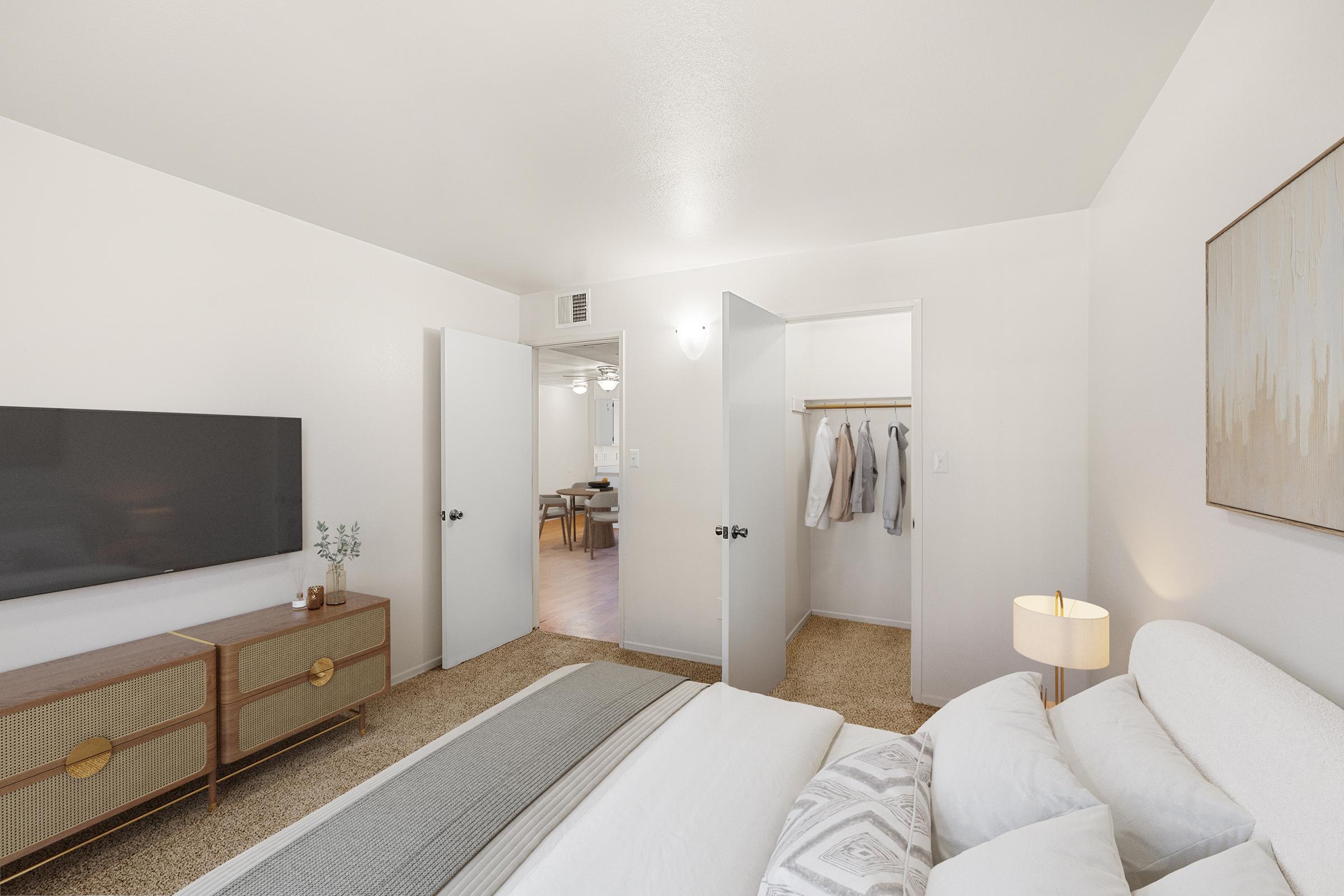 A cozy bedroom featuring a comfortable bed with a gray blanket, a stylish nightstand with a lamp, and a modern television on a wooden console. Two open white doors lead to a closet, and there is a glimpse of a dining area with a ceiling fan in the background. The walls are painted in light colors, enhancing the spacious feel.