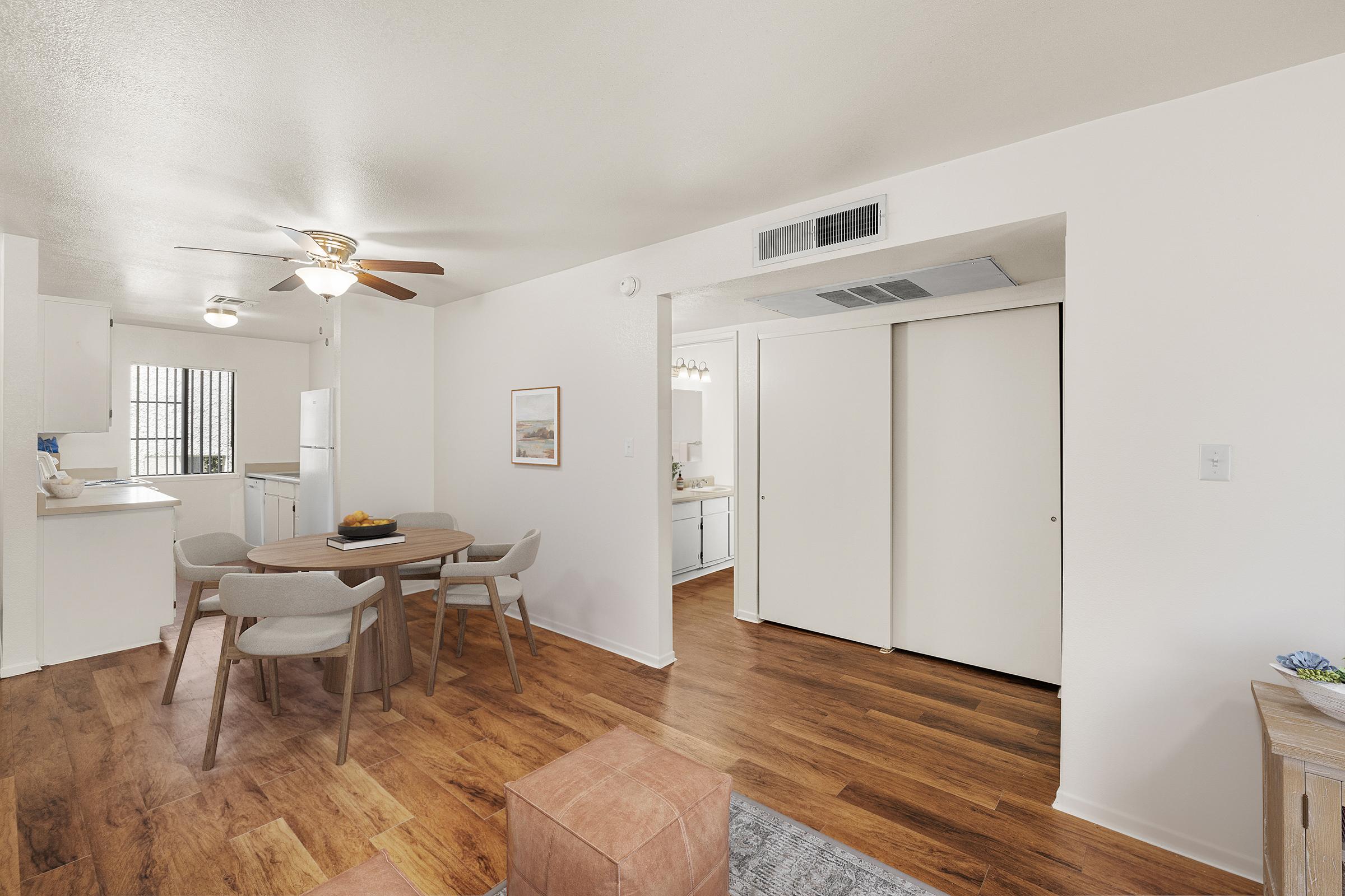 A bright and spacious living area featuring a round dining table with four chairs, a ceiling fan, and hardwood flooring. There is a doorway leading to a kitchen area visible in the background, with white cabinetry. A cozy ottoman is placed on the floor, and a framed picture adds a decorative touch to the wall.