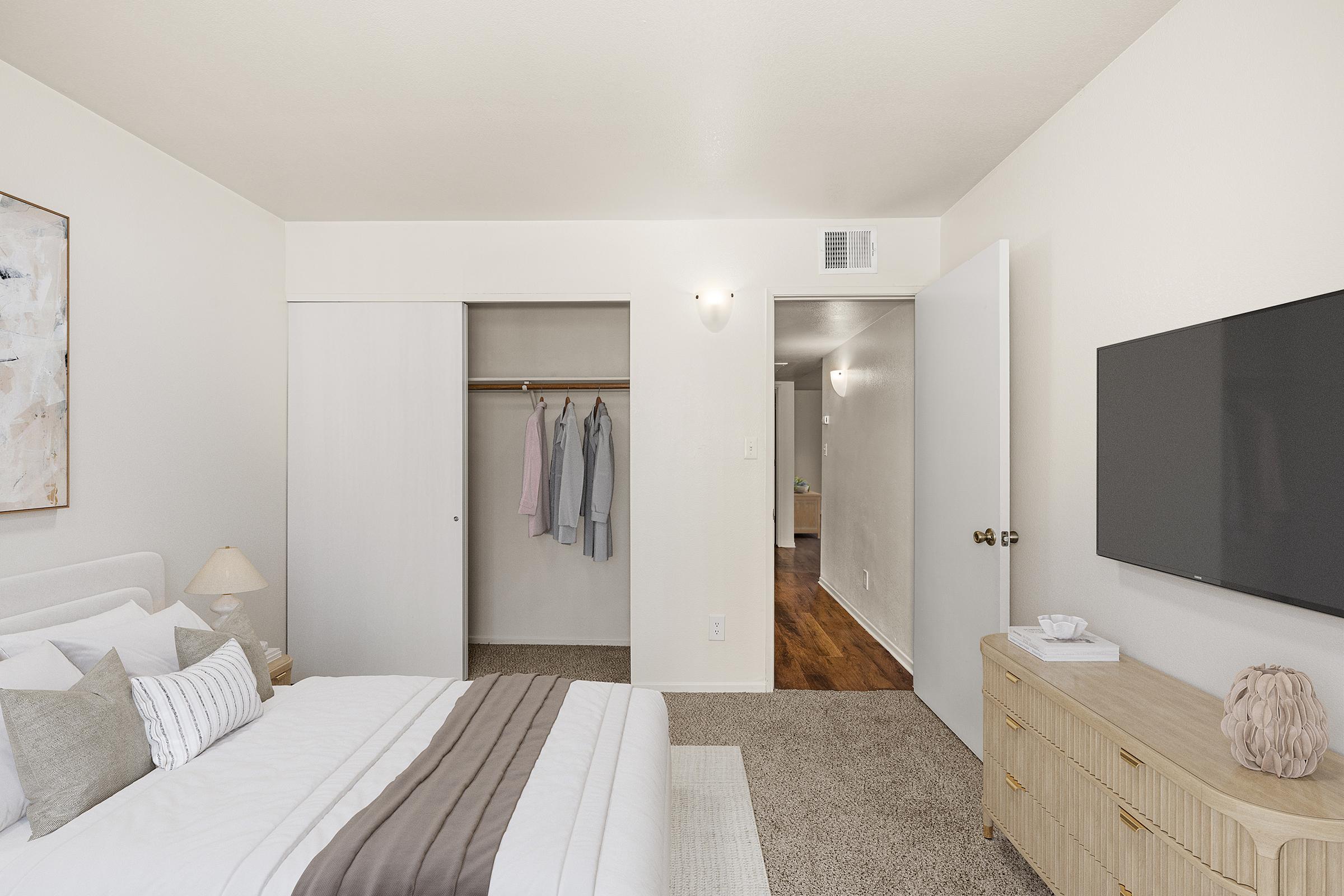 A cozy bedroom featuring a neatly made bed with decorative pillows, a bedside lamp, and a wooden dresser. A closet with hanging clothes is visible, along with a TV mounted on the wall. The room has neutral-colored walls and carpet, creating a calm atmosphere, and a door leading to another room.