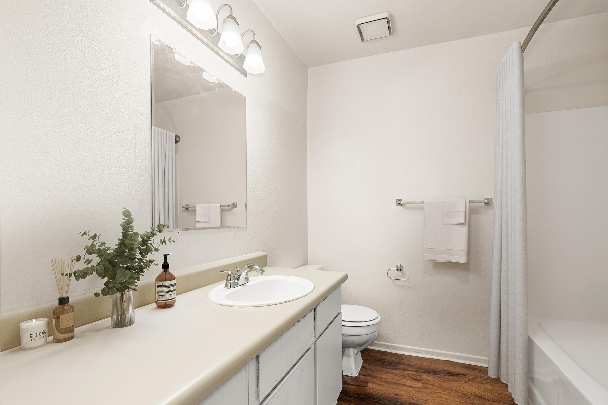 Bright, modern bathroom featuring a beige countertop sink, a large mirror, and white cabinetry. There is a white shower curtain and a toilet nearby. Decor includes a small plant and candles on the counter, along with neatly hung towels on a bar. The flooring is wood, contributing to a clean and inviting atmosphere.