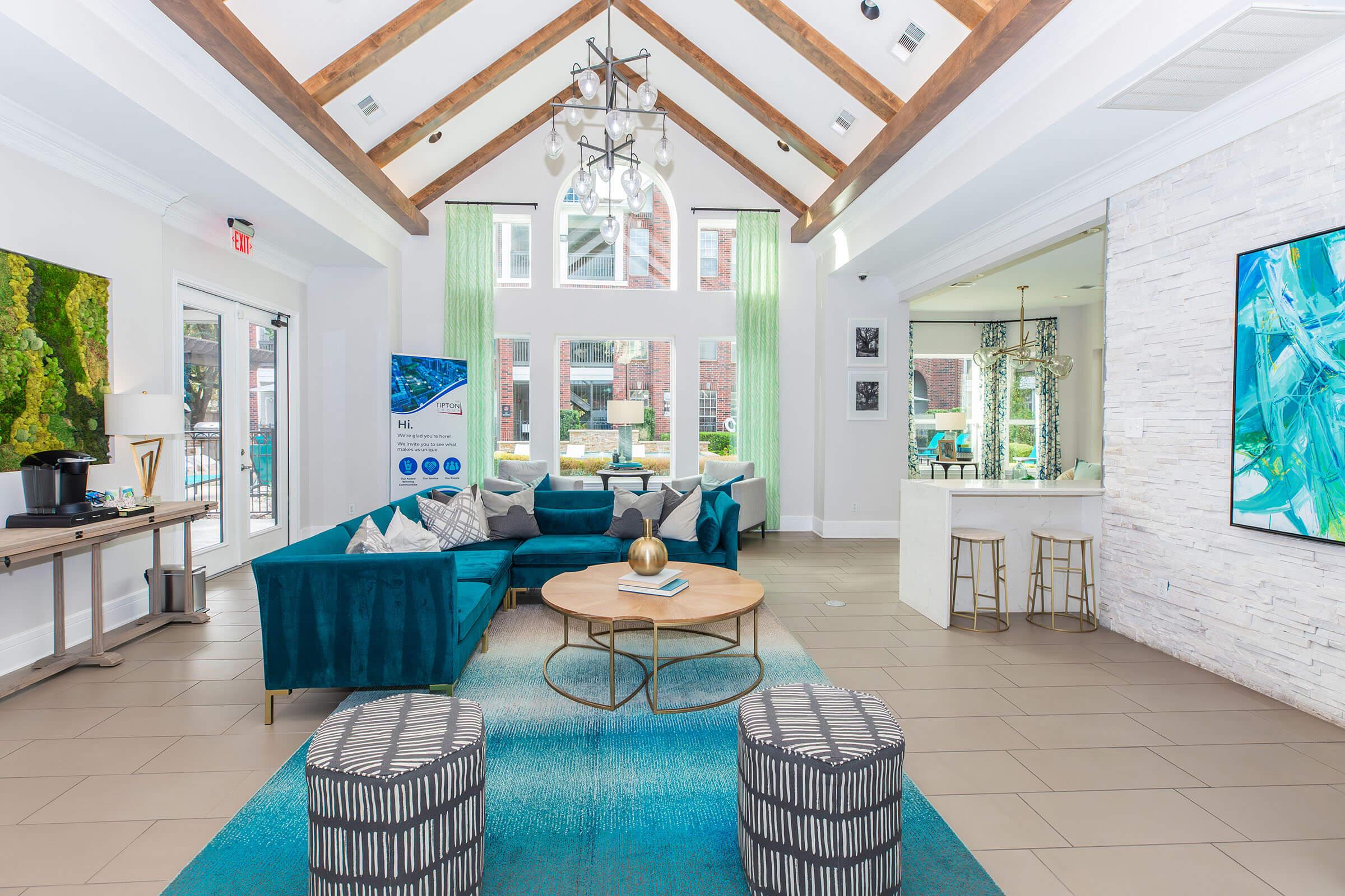 A spacious, modern lounge area featuring high ceilings with wooden beams, large windows, and bright green curtains. The room is furnished with a teal sofa, a round coffee table, and patterned stools on a blue area rug. A sleek bar area is visible in the background, creating a welcoming and stylish atmosphere.